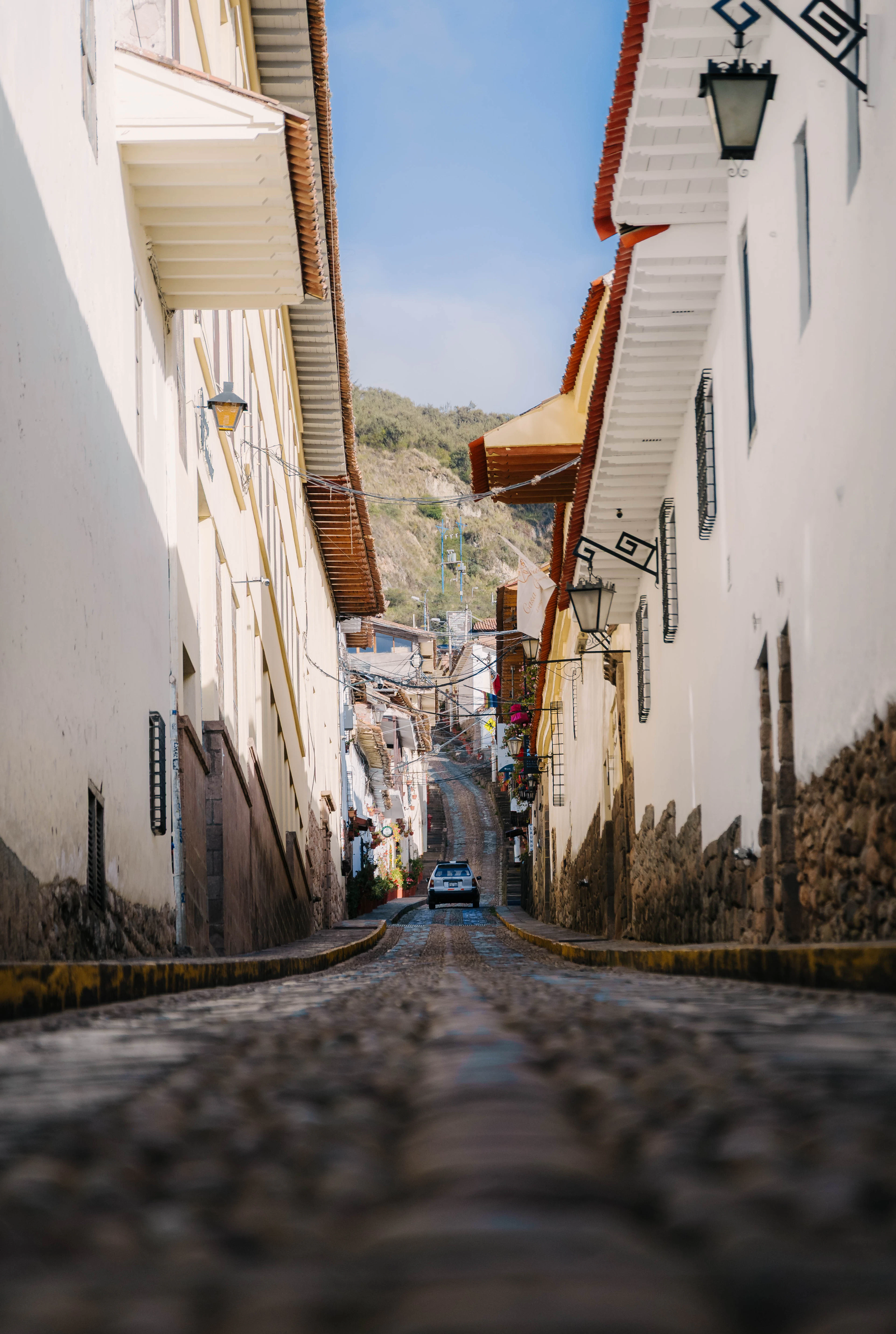 A narrow cobbled street on Cusco's upper slopes seen from pavement level, whitewashed colonial houses with red-tile eaves and wrought-iron lanterns closing in on both sides, a green hillside at the top of the climb.