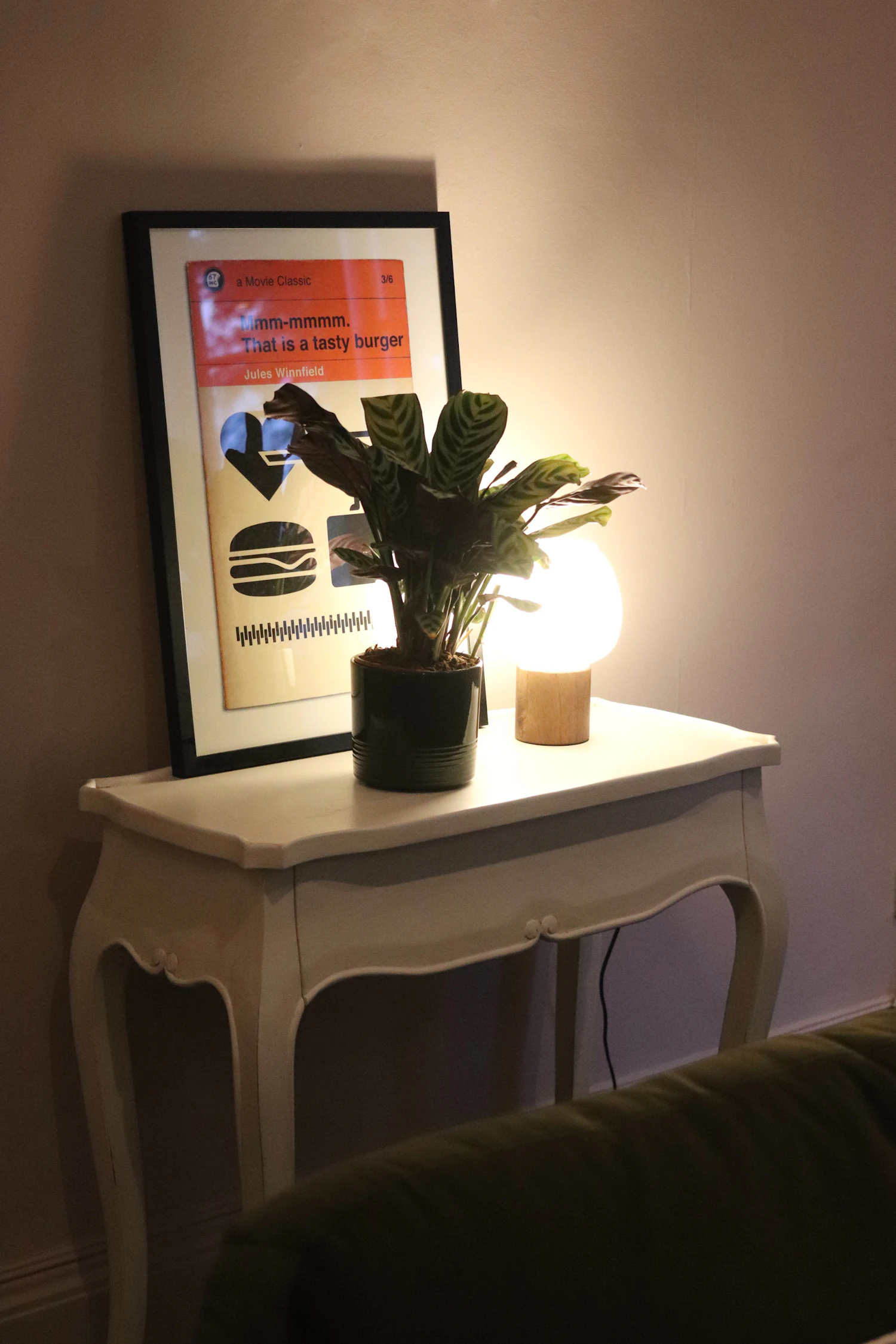 A framed print on a small side table next to a lit lamp and a plant