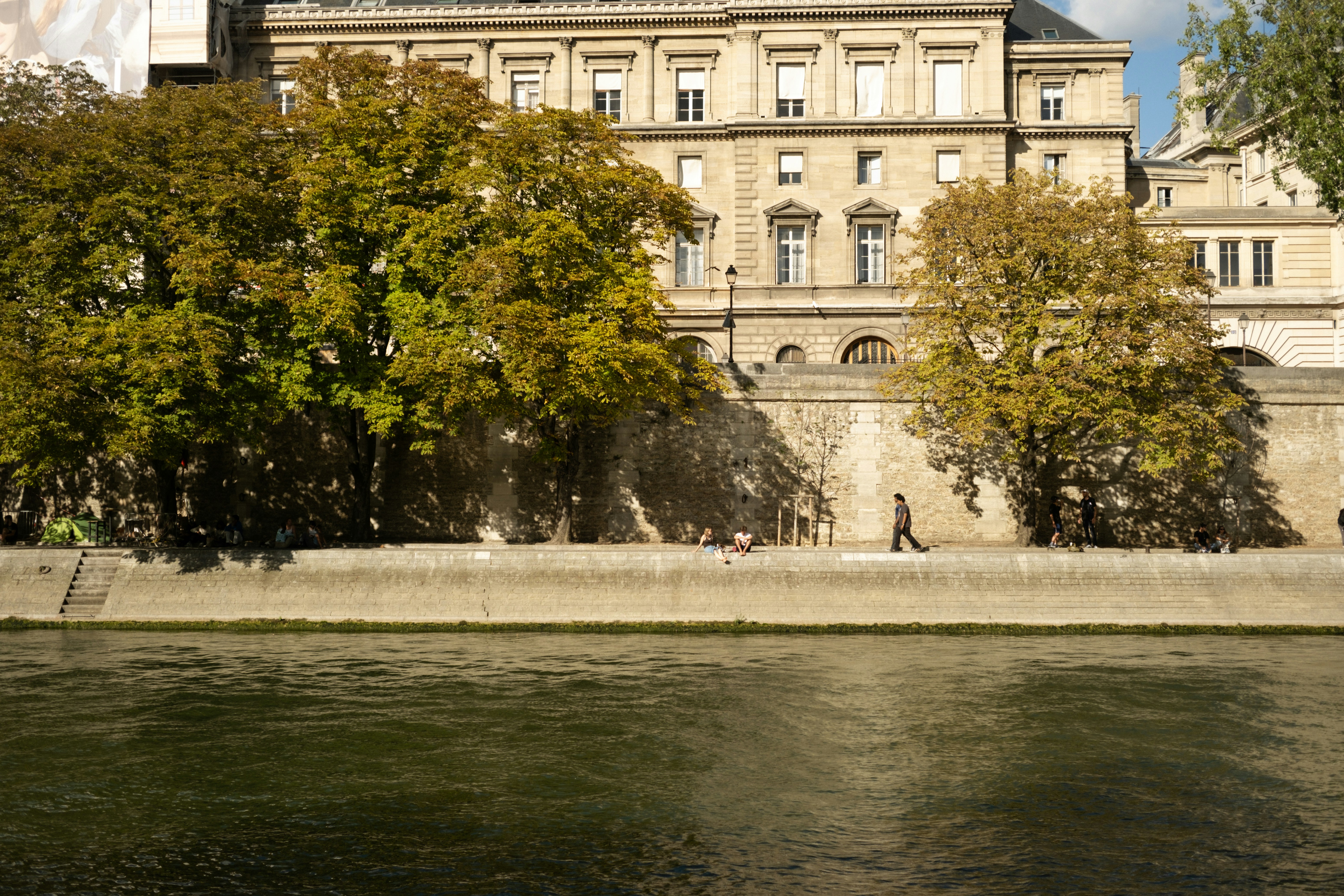 A person walks along a river with trees and buildings.