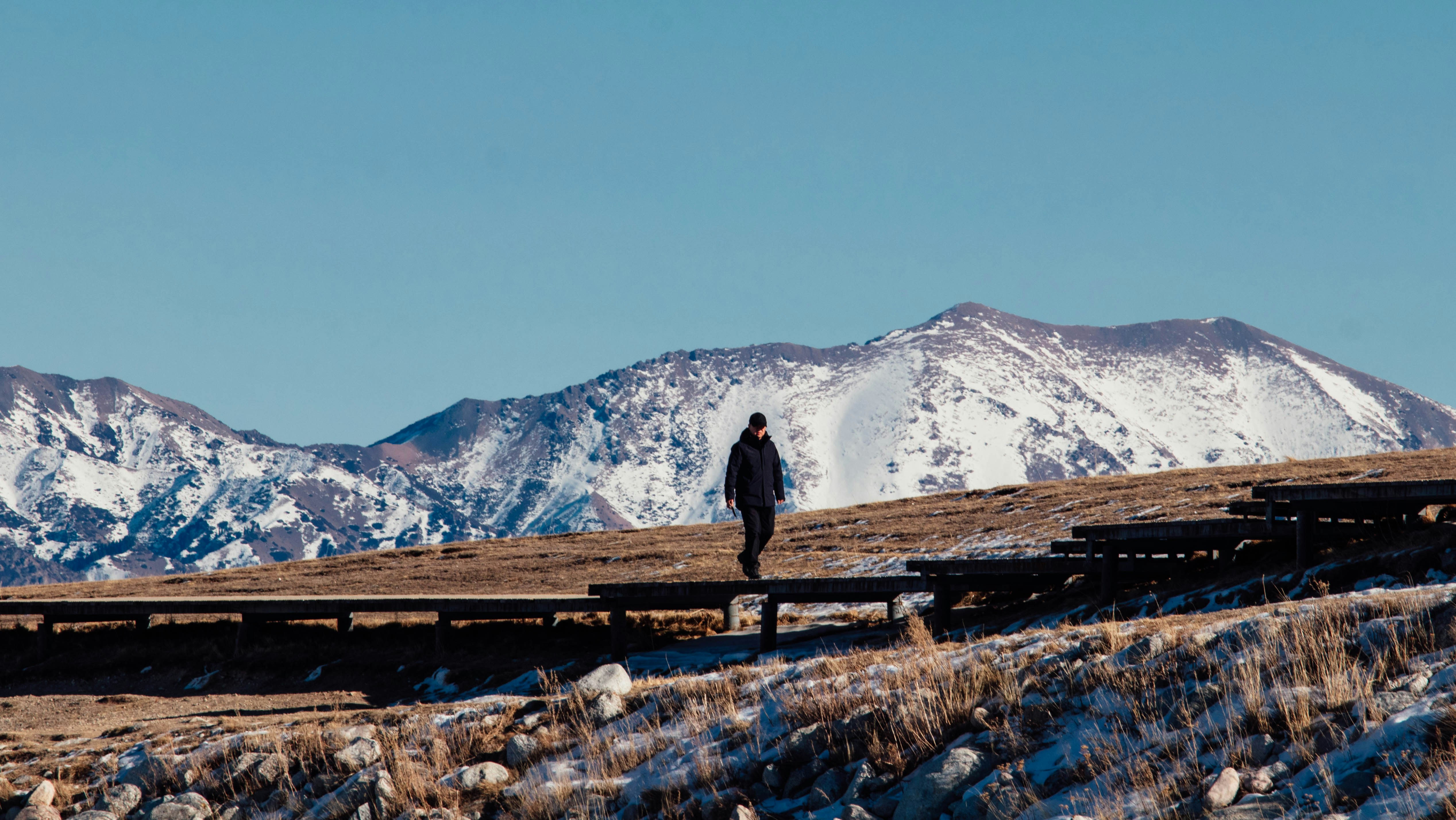 A person walks on a wooden path towards snowy mountains.