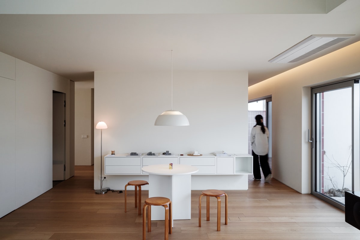 Ambiente minimalista ordenado y despejado, con piso de madera clara, aparador blanco sin objetos encima, mesa redonda y banquetas de madera