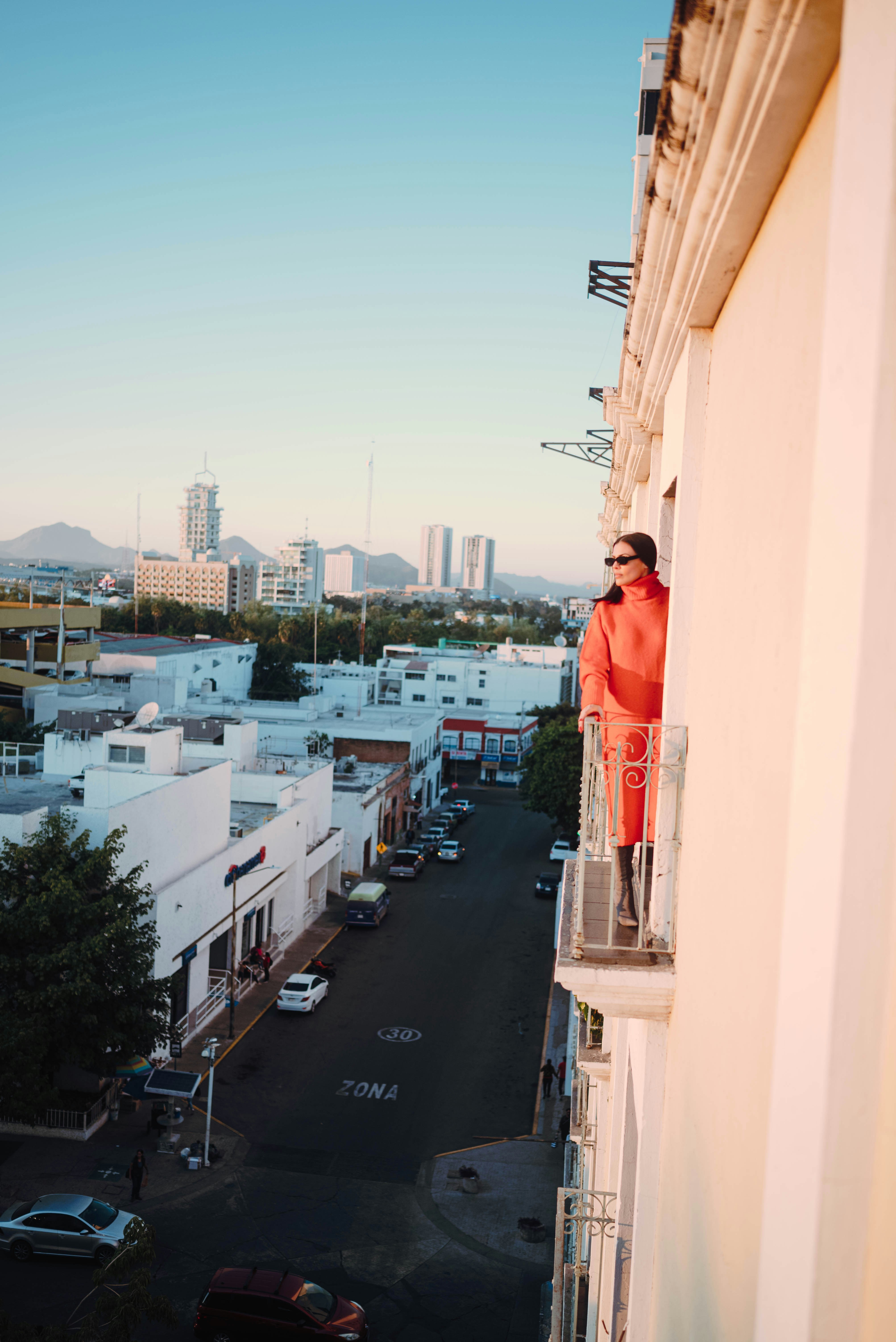 Woman in orange coat on balcony overlooking city street.