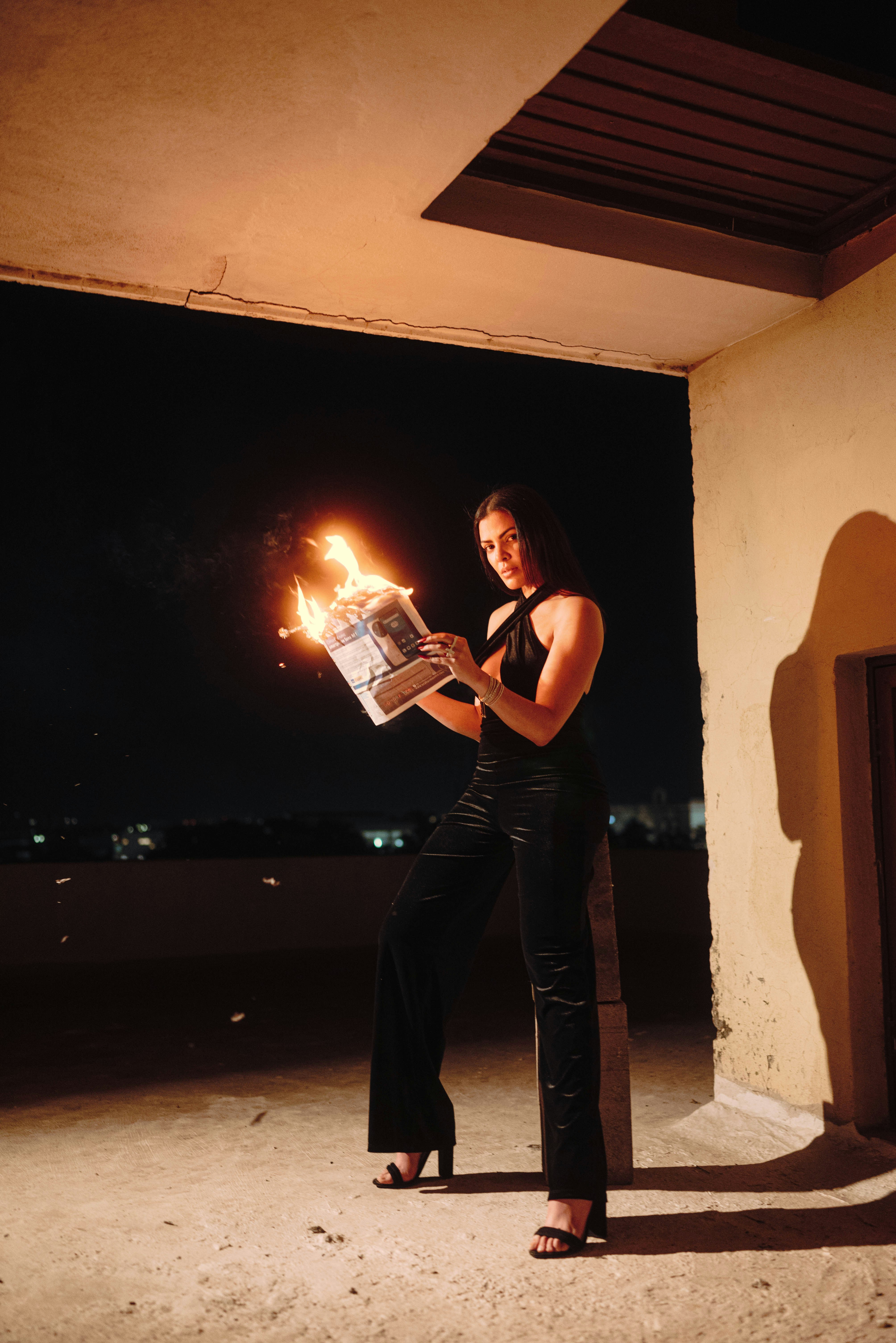 Woman in black holding a burning newspaper at night.