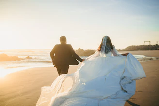 Newlyweds run hand in hand on a beach at sunset.