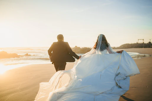 Newlyweds run hand in hand on a beach at sunset.