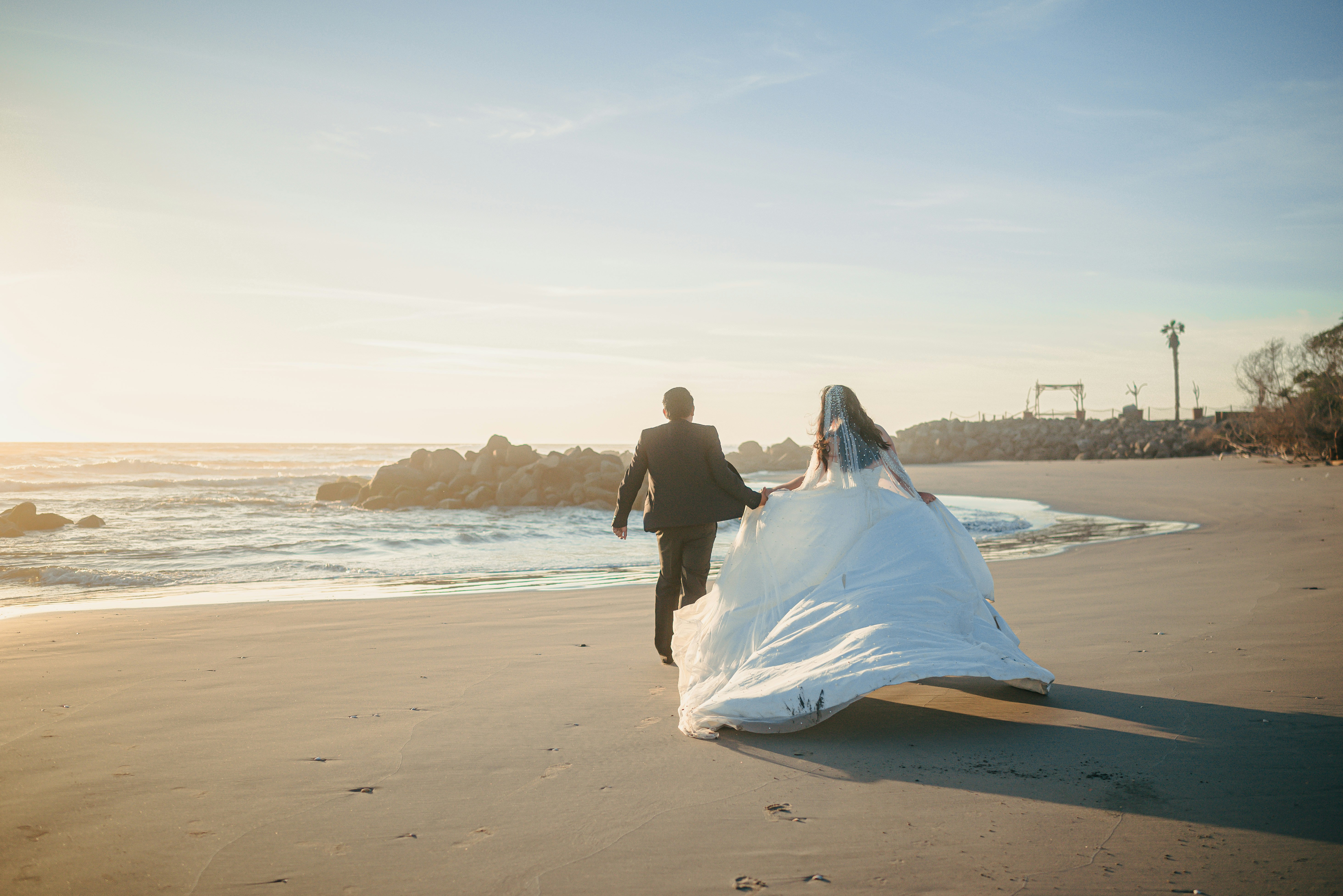 Recém-casados correndo numa praia ao pôr do sol.