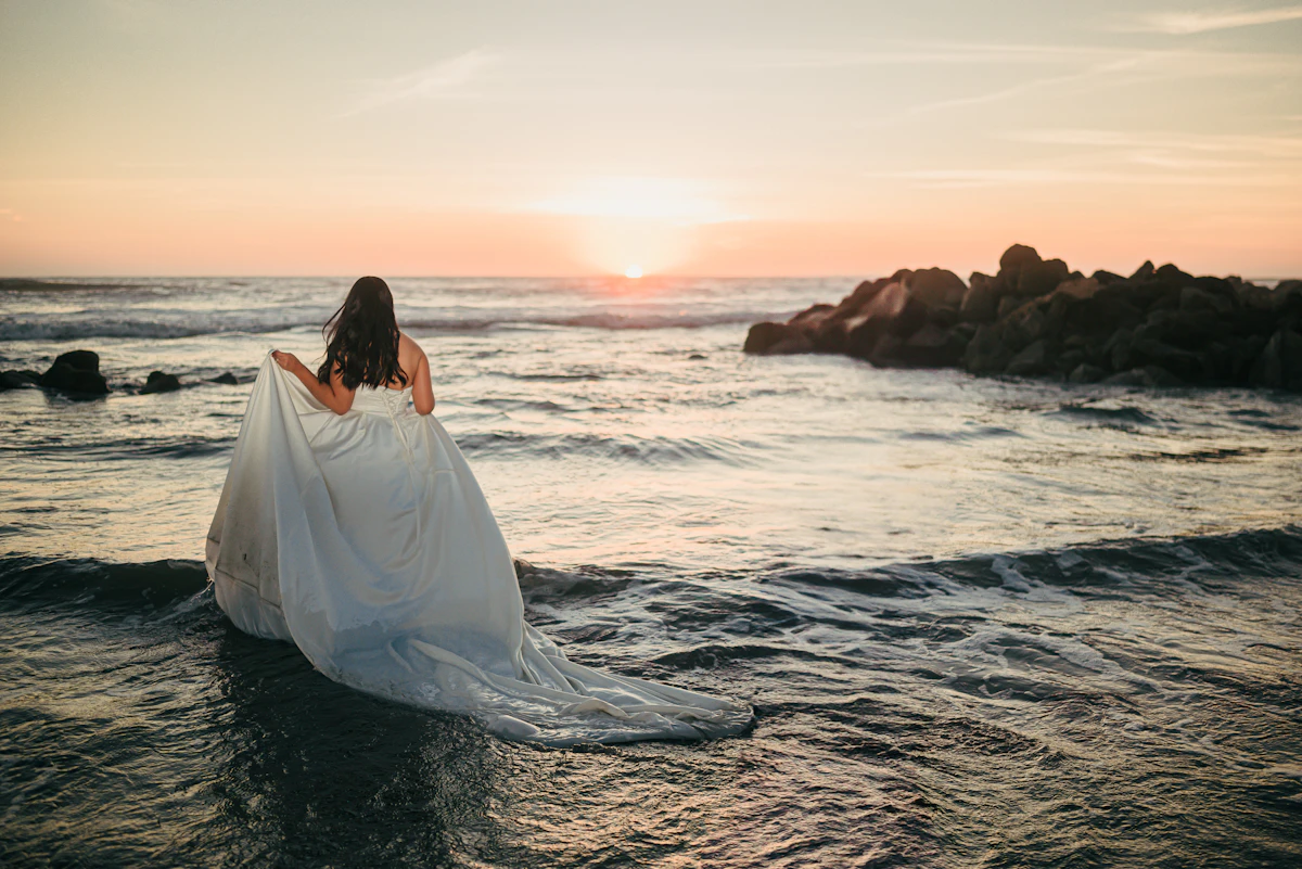 A bride in a flowing white gown walking into the ocean at golden hour
