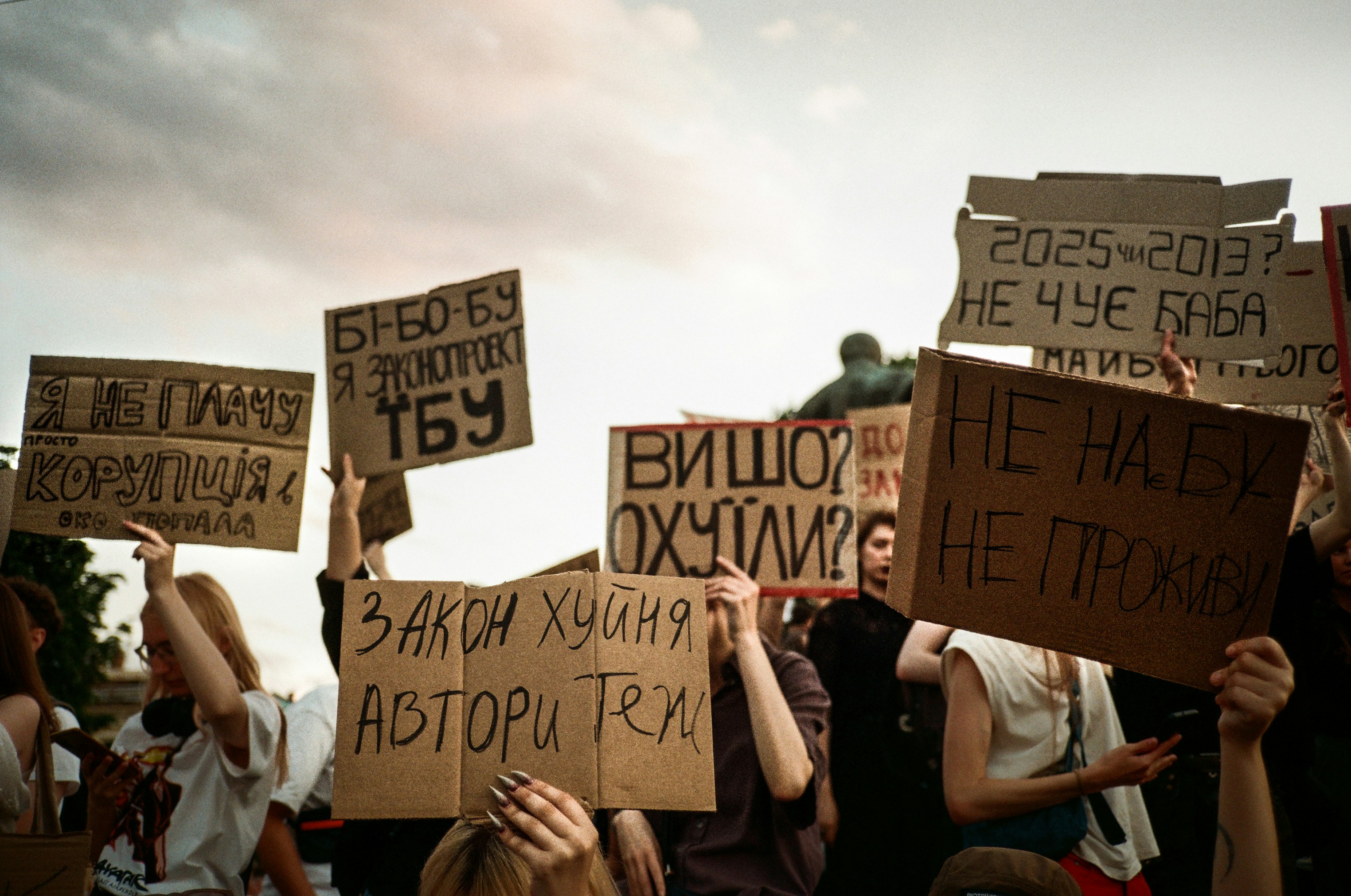 Protesters hold signs with handwritten messages against a cloudy sky.