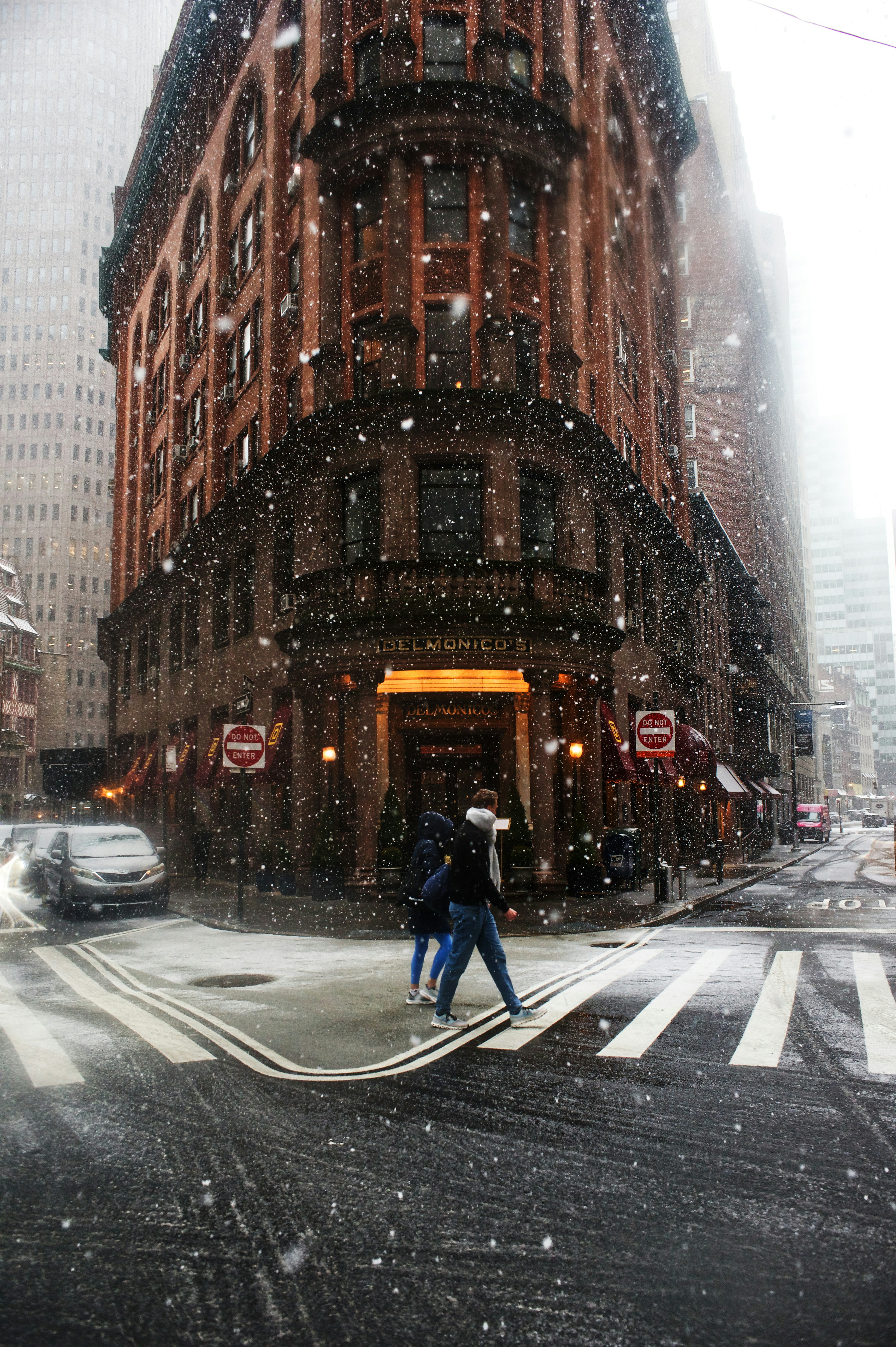 People cross street in front of building during snowfall
