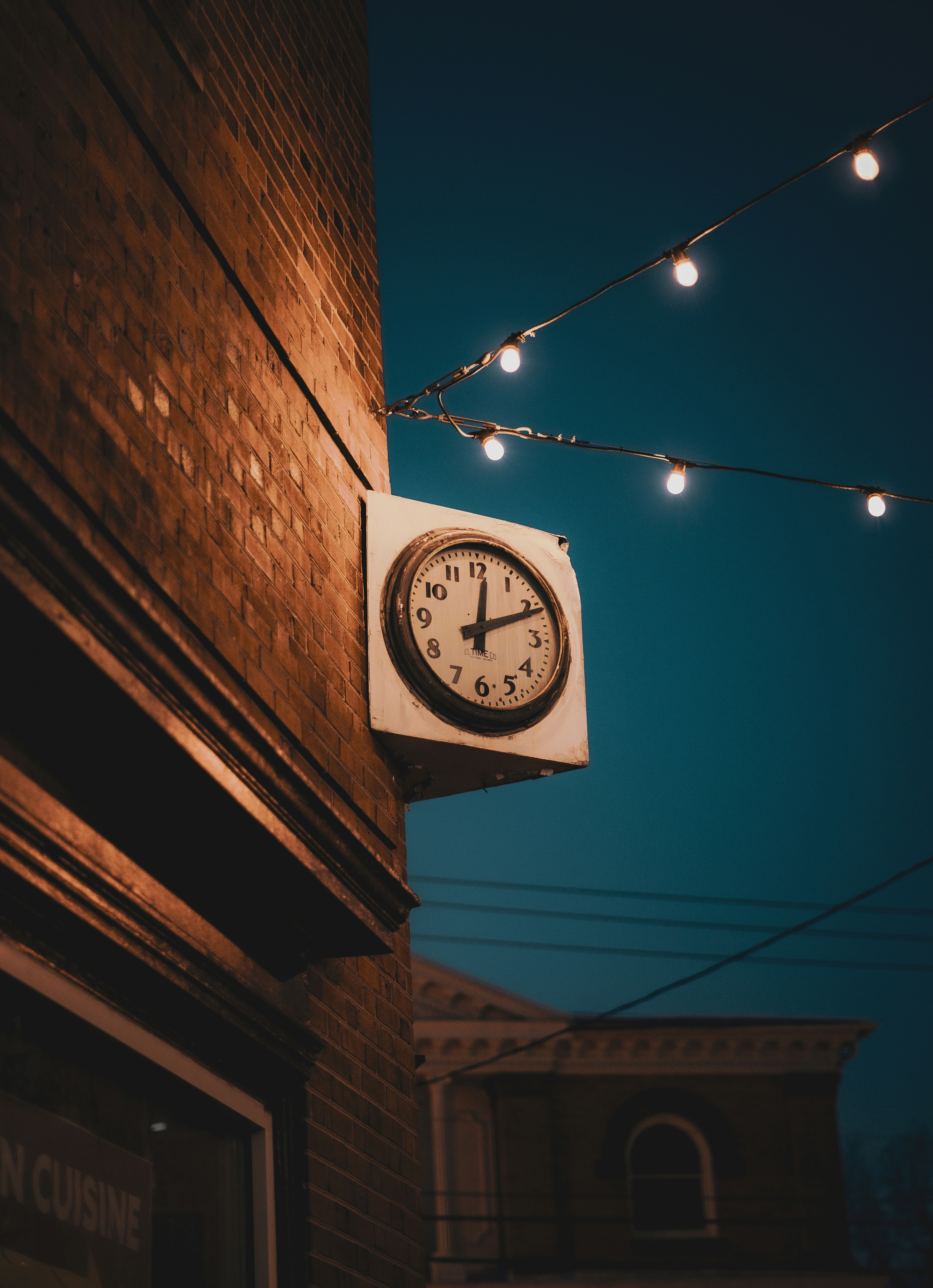 A clock hangs on a brick wall with string lights.