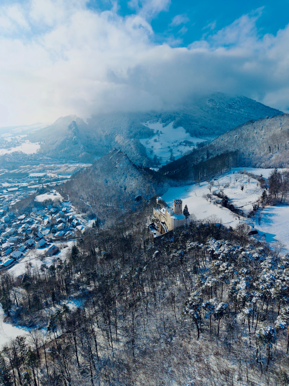 The image presents an aerial view of a mountainous landscape blanketed with snow, featuring a castle-like structure nestled among the trees and hills. The sky above is a clear blue, dotted with fluffy white clouds that add depth to the scene. The perspective from above provides a comprehensive look at the natural beauty of this winter wonderland.