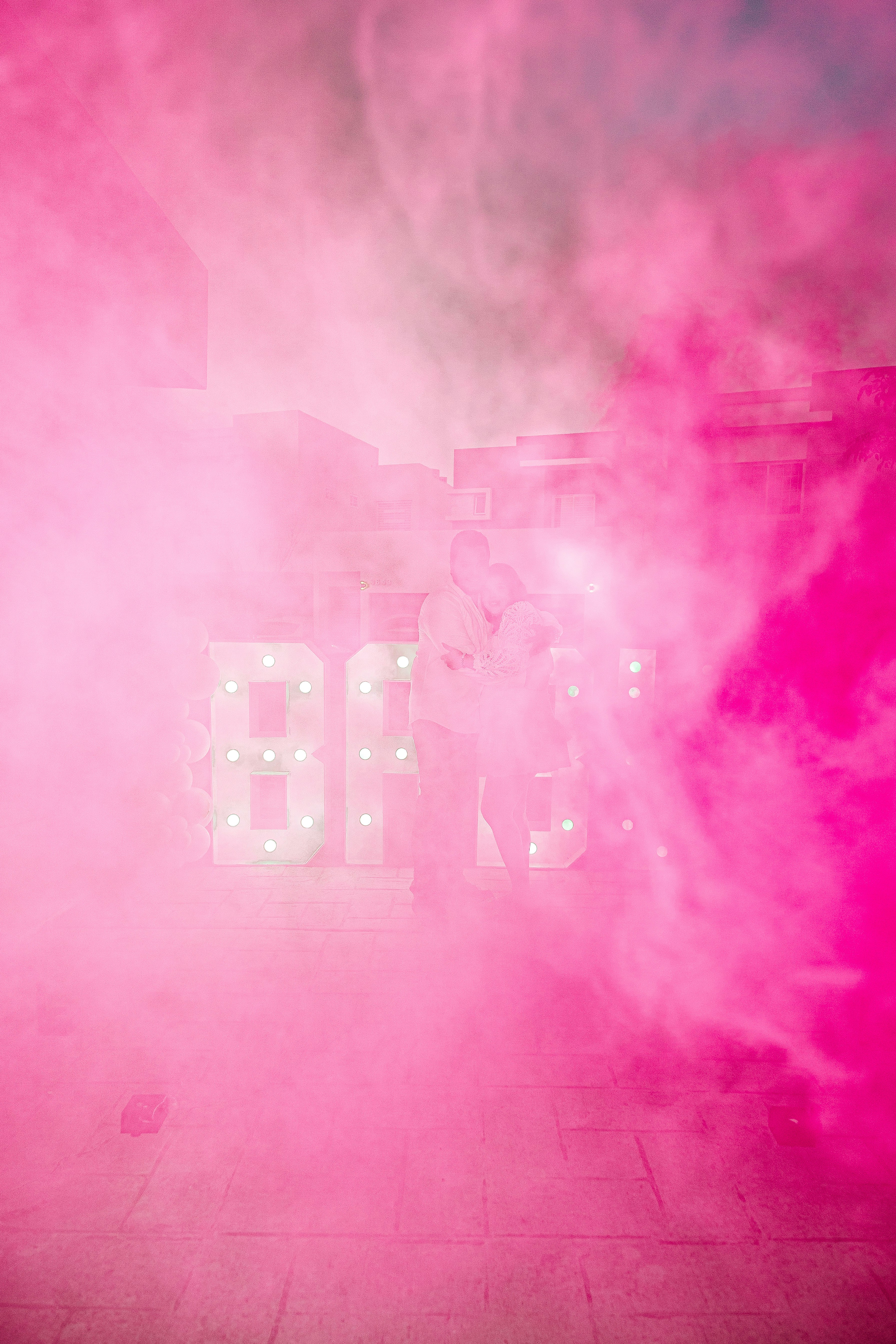Couple kissing behind bright pink smoke and lit letters.