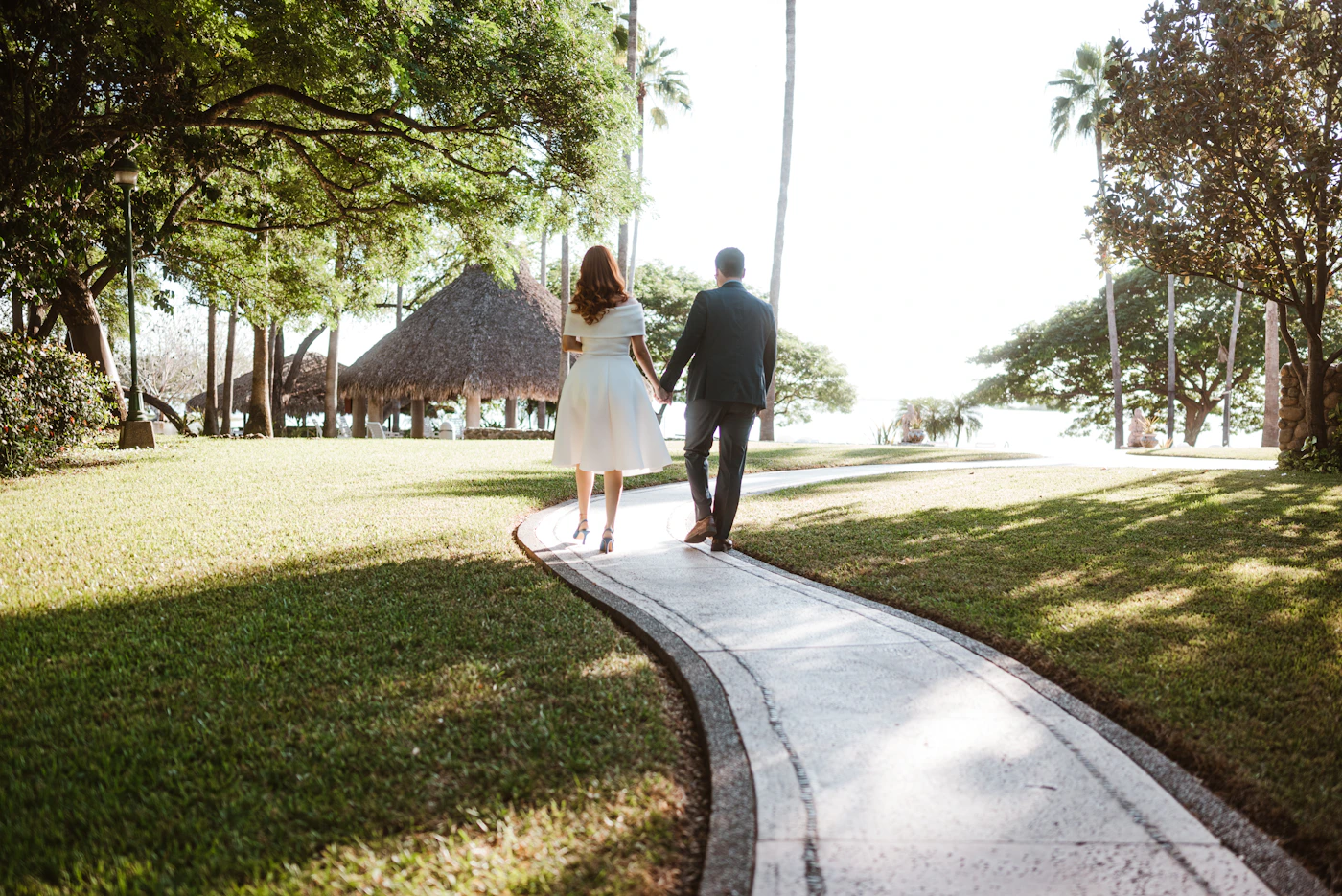 Couple in wedding attire walking hand in hand on a palm-lined path