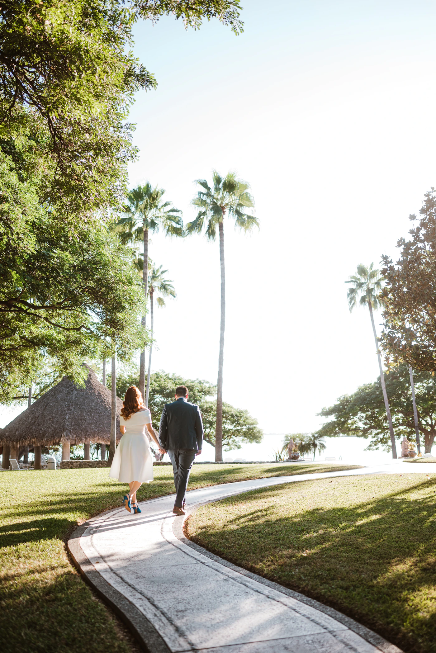 Couple in wedding attire walking a palm-lined path toward the ocean