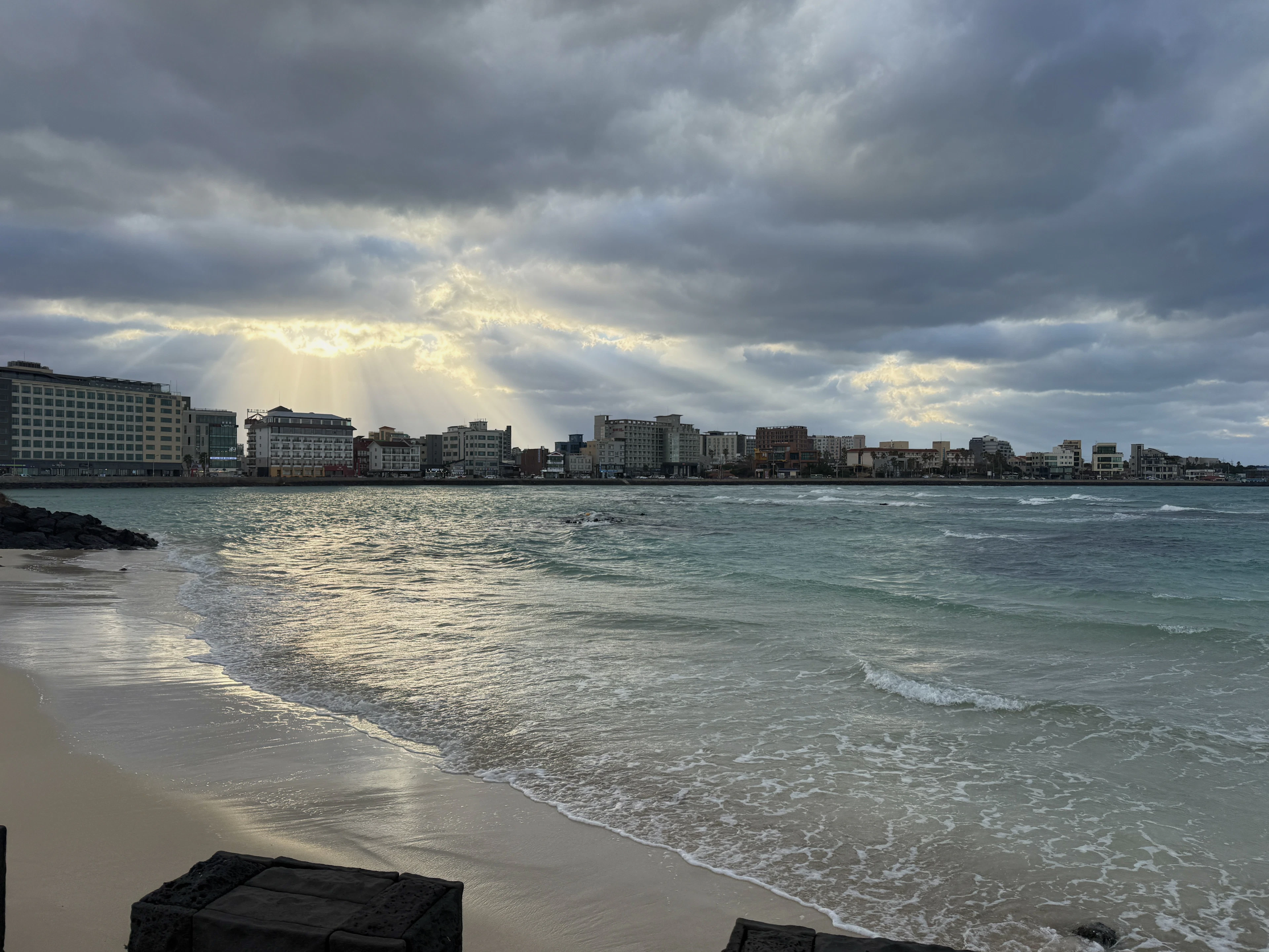 Sun rays break through clouds over a coastal city skyline seen from a sandy beach.