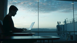 Man working on laptop by airport window at sunrise