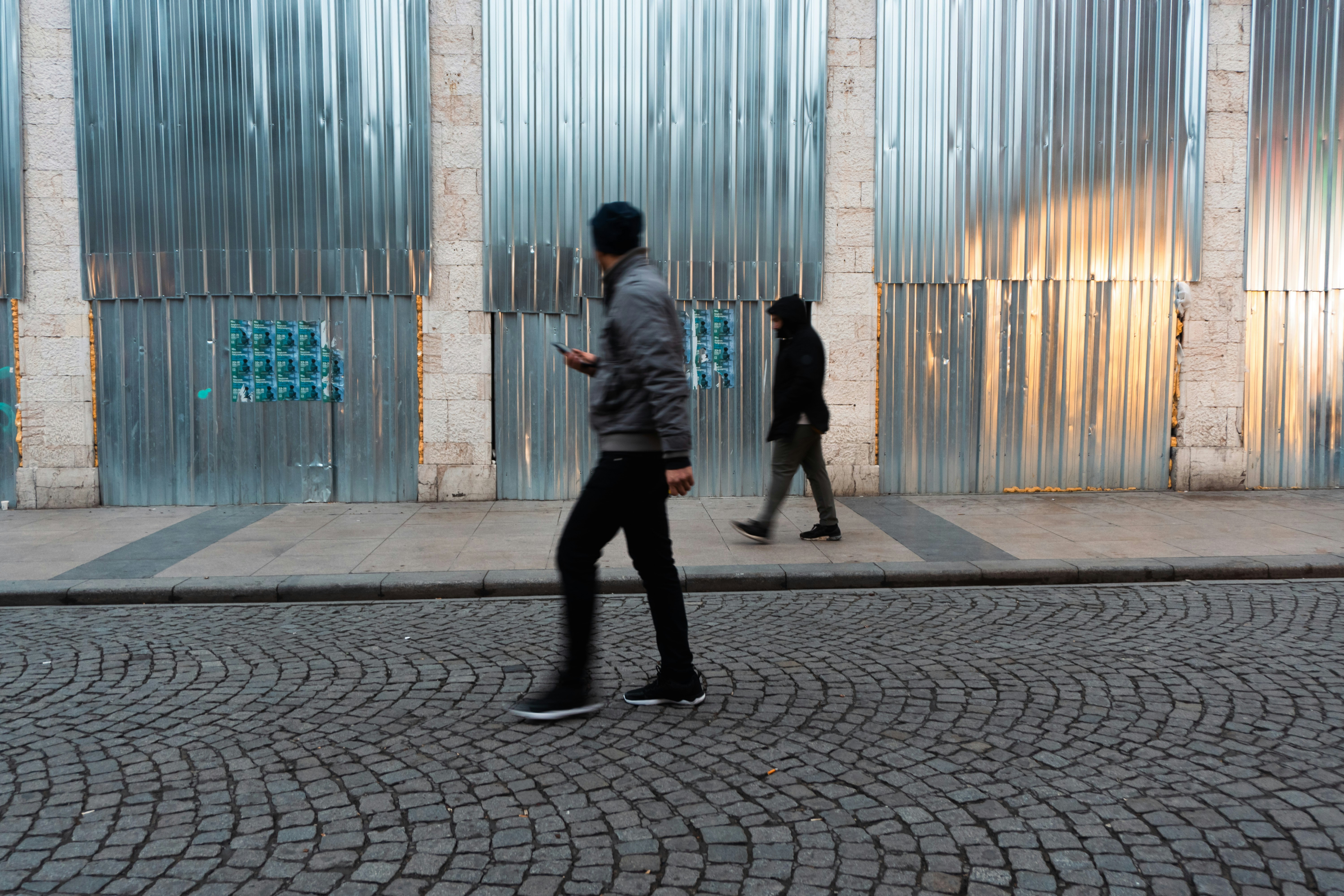 Two people walk past a corrugated metal wall.
