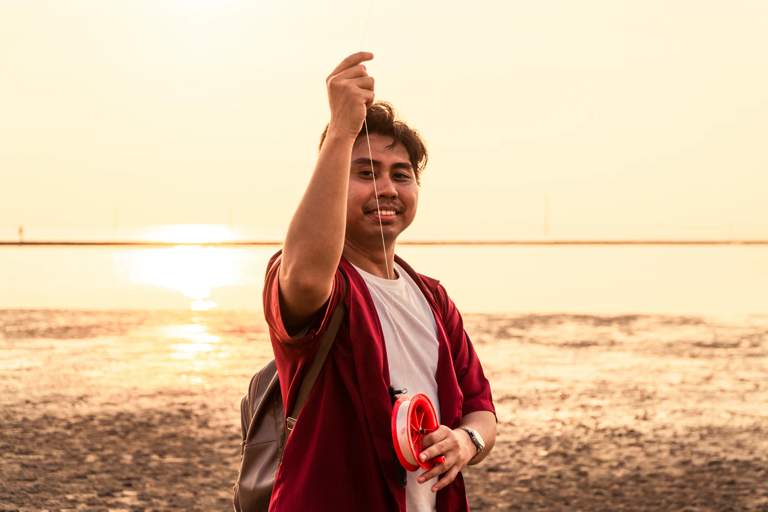 A young man on a beach at sunset holding up his kite line and reel