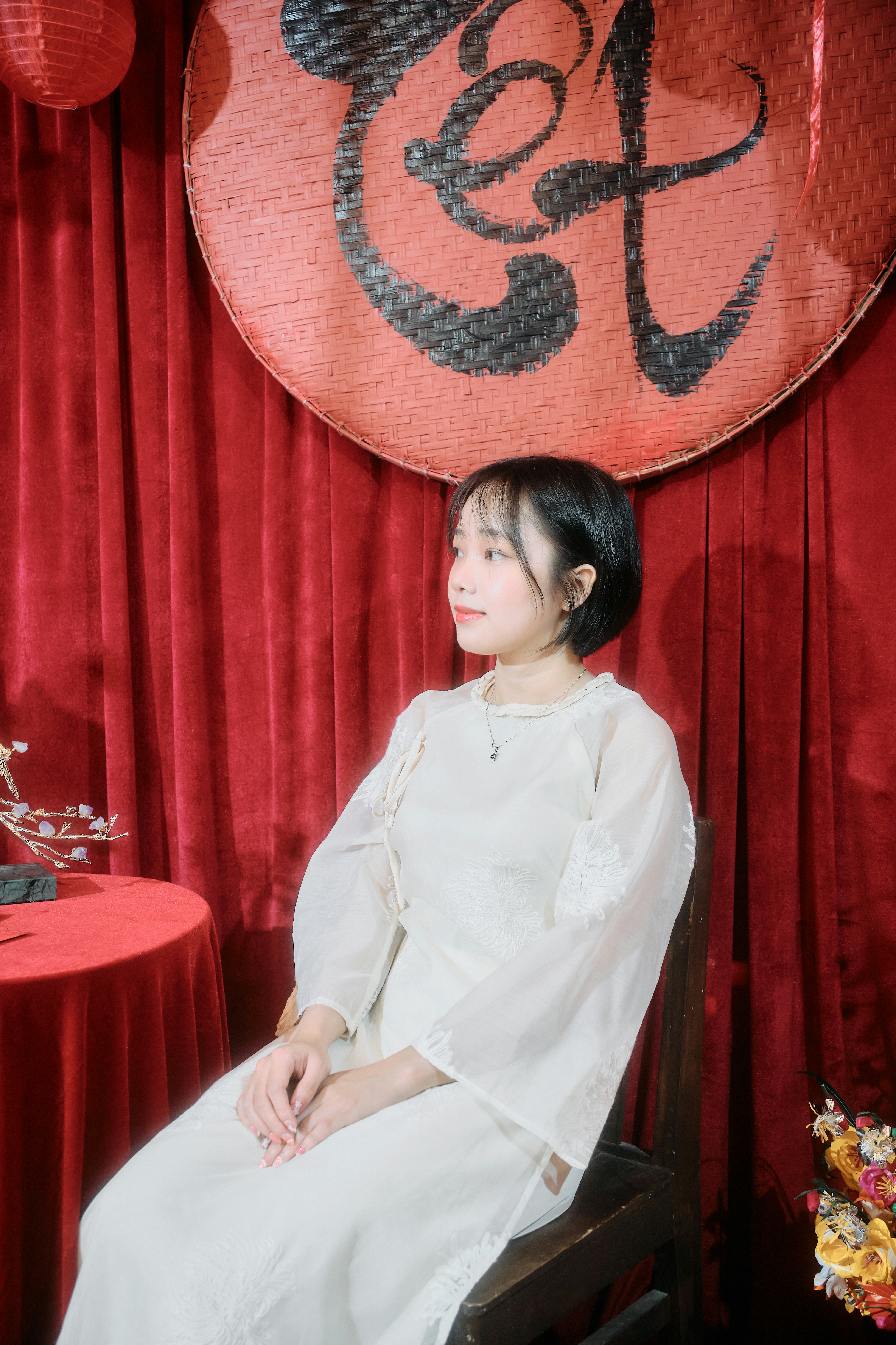 Young woman in white dress sits before red curtains.