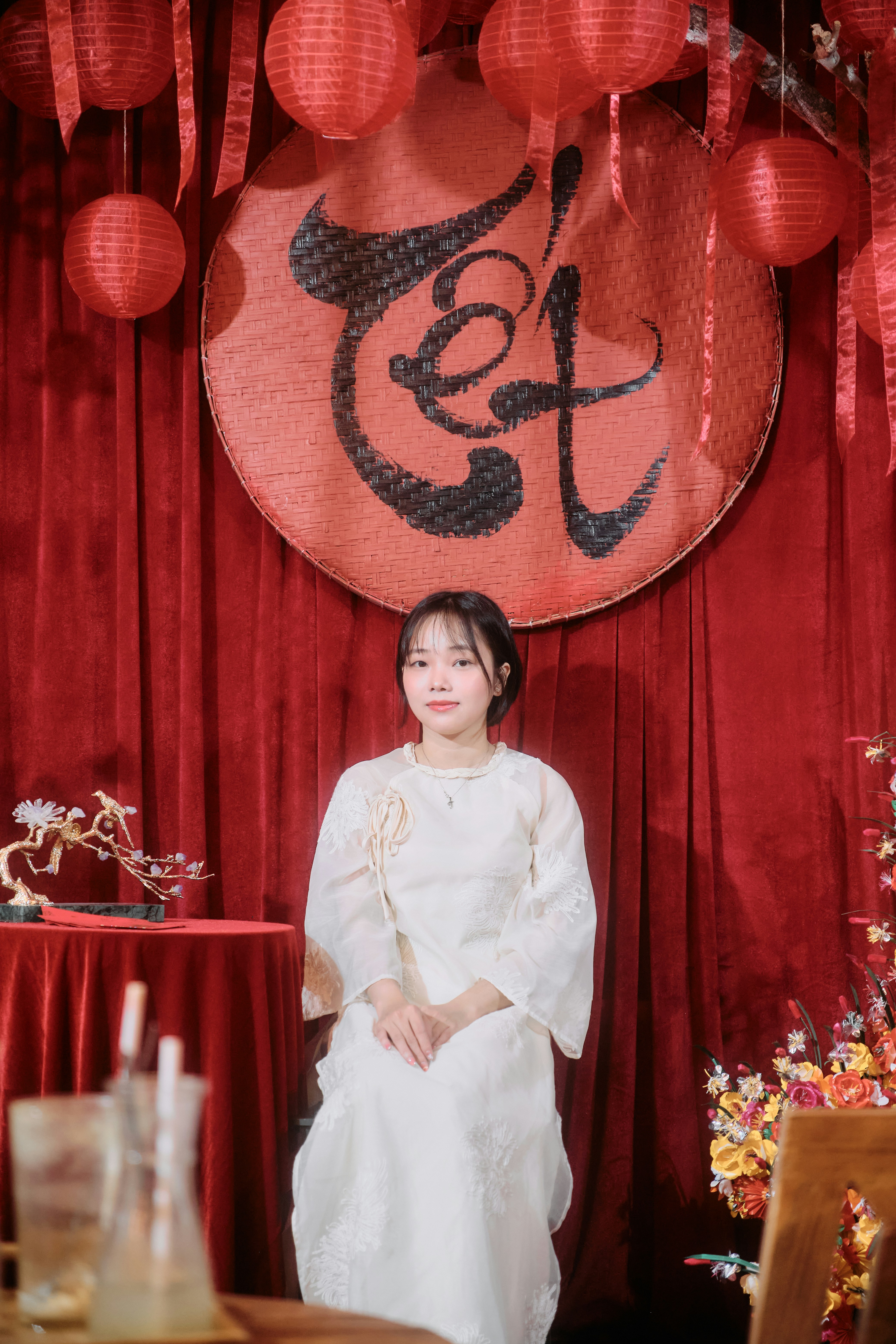 A young woman in traditional attire sits before tet decorations.