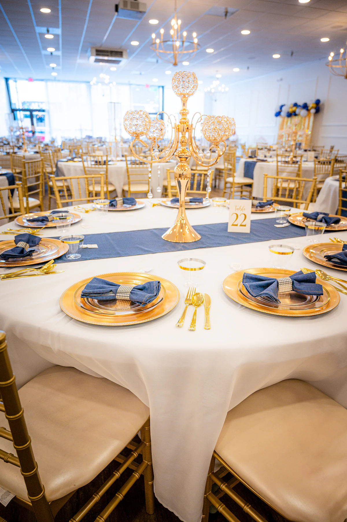 An elegant banquet table with gold chairs, gold charger plates, navy napkins and a gold centerpiece