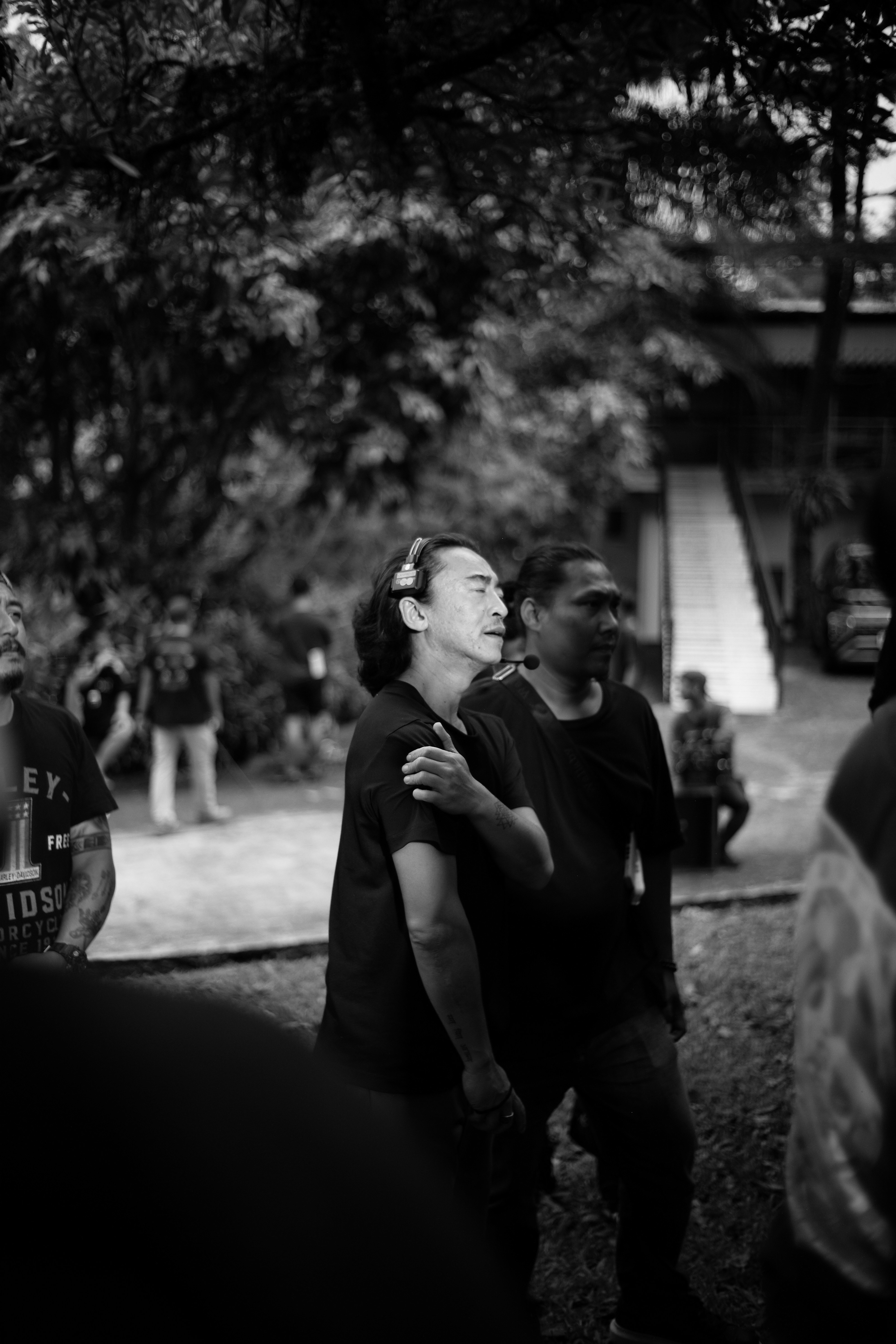 Two men standing together outdoors with trees and stairs.