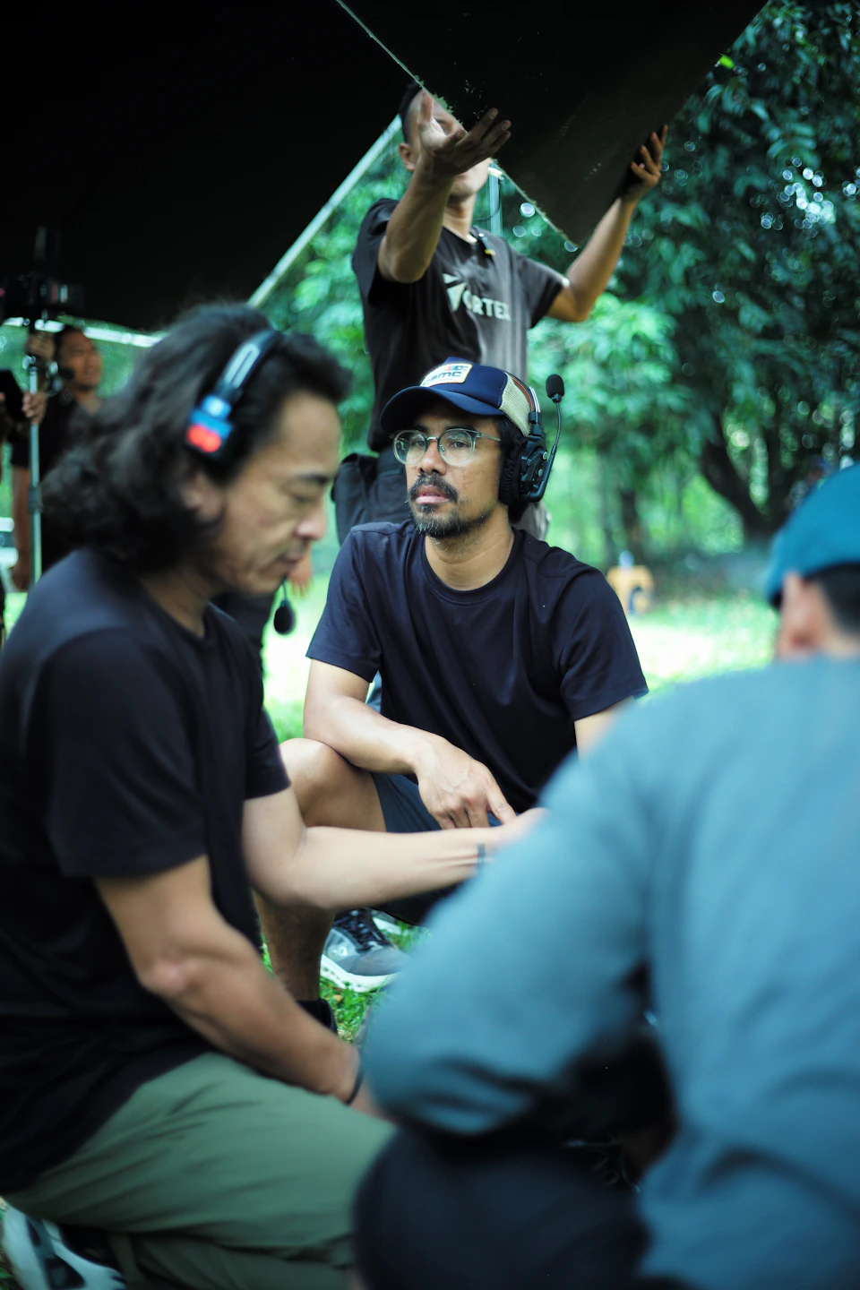 The image shows a group of people gathered under a large black tarp, likely on a film set or outdoor shoot location. The individuals are engaged in conversation and appear to be enjoying each other's company. One person is wearing a baseball cap, while another has a backpack nearby. The scene suggests that they might be discussing the day's filming activities or sharing stories about their experiences.