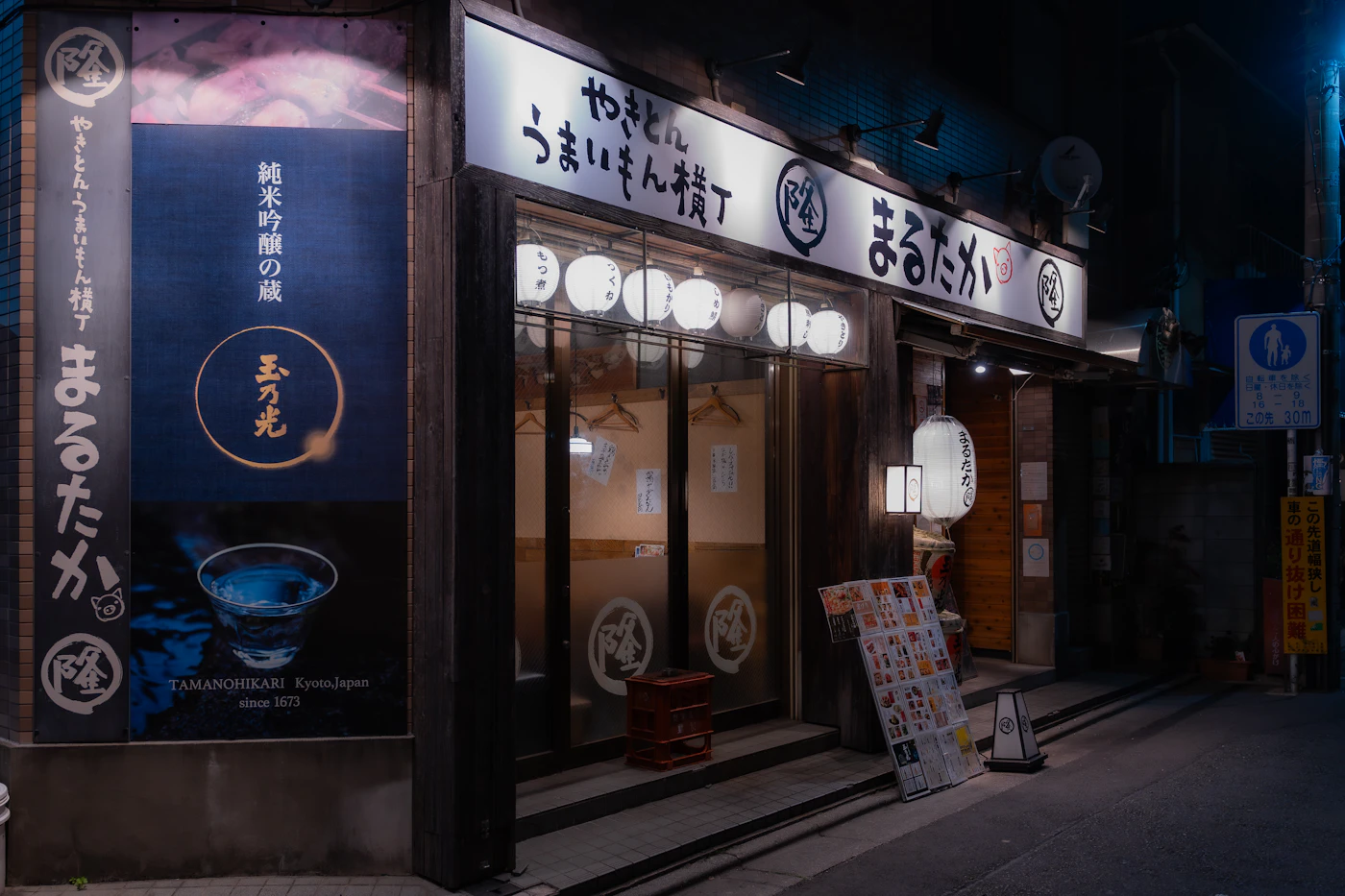 A lantern-lit izakaya alley in Japan at night, with a menu stand out front and a pedestrian sign on the corner.