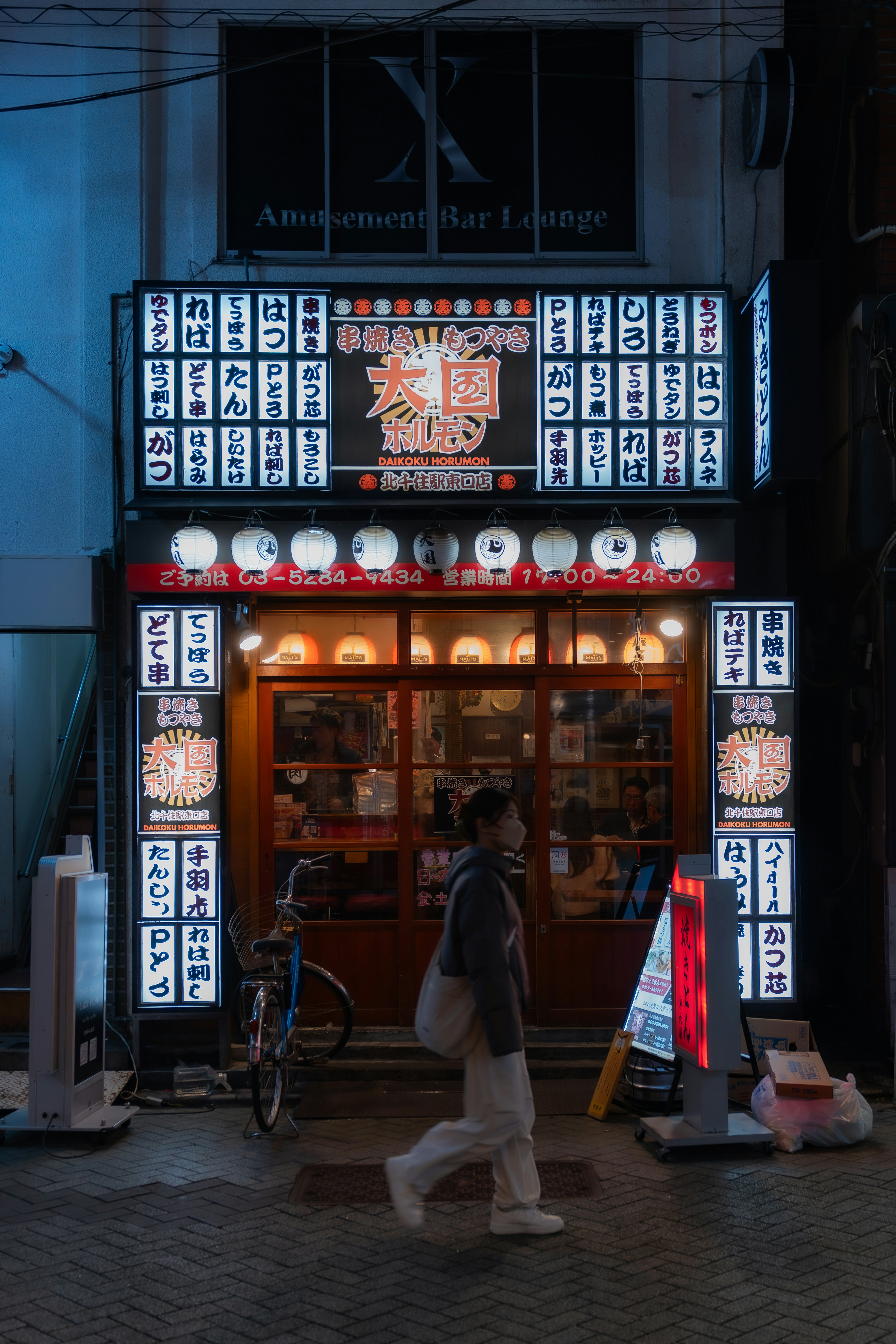 Person walks past a brightly lit japanese restaurant at night.
