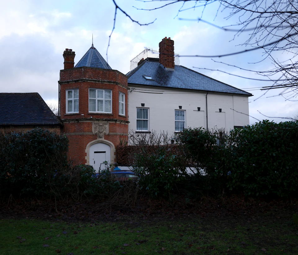The image depicts a large, two-story brick house with a white exterior and a blue roof, situated on a grassy lawn. The house features a chimney on one side and a small porch with a white railing. In front of the house, there is a small garden with bushes and trees.