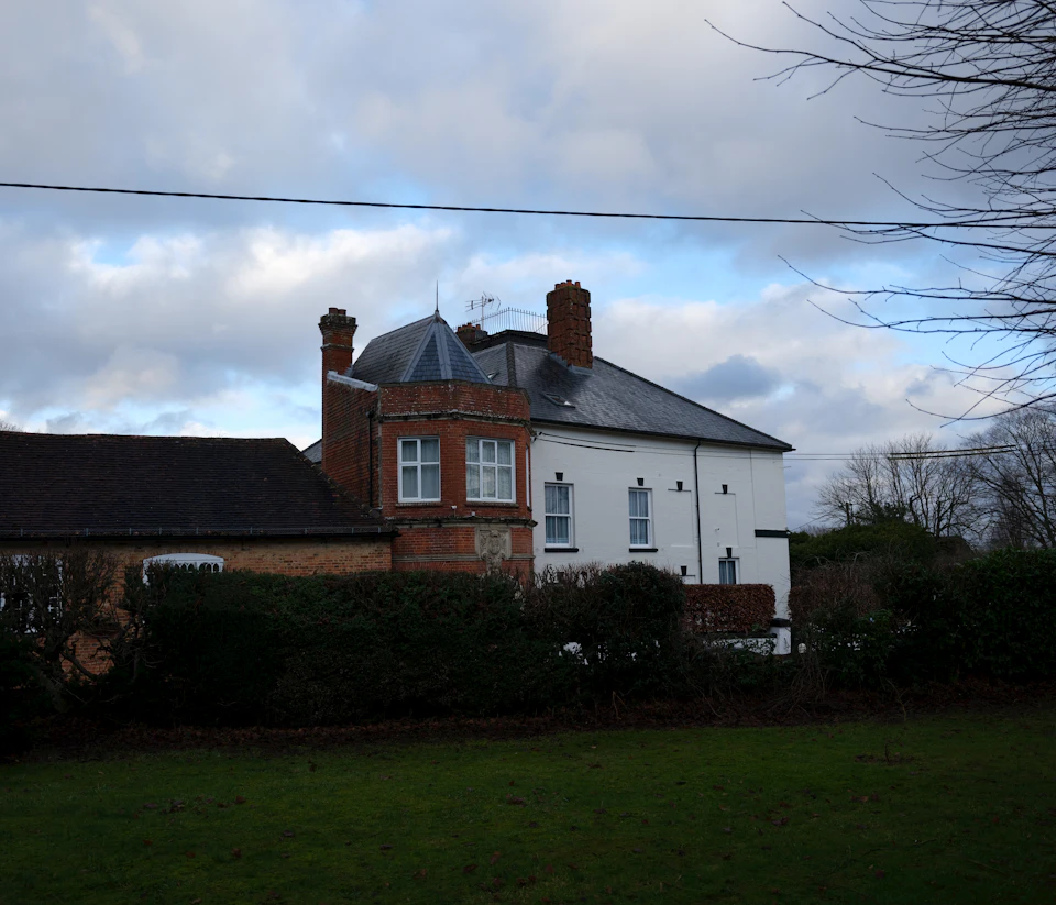 The image depicts a two-story brick house with a white roof, situated on a grassy lawn. The house features a chimney and a small tower on its left side. A telephone pole stands tall next to the house, and a tree can be seen in front of it. In the background, there are trees that add depth to the scene.