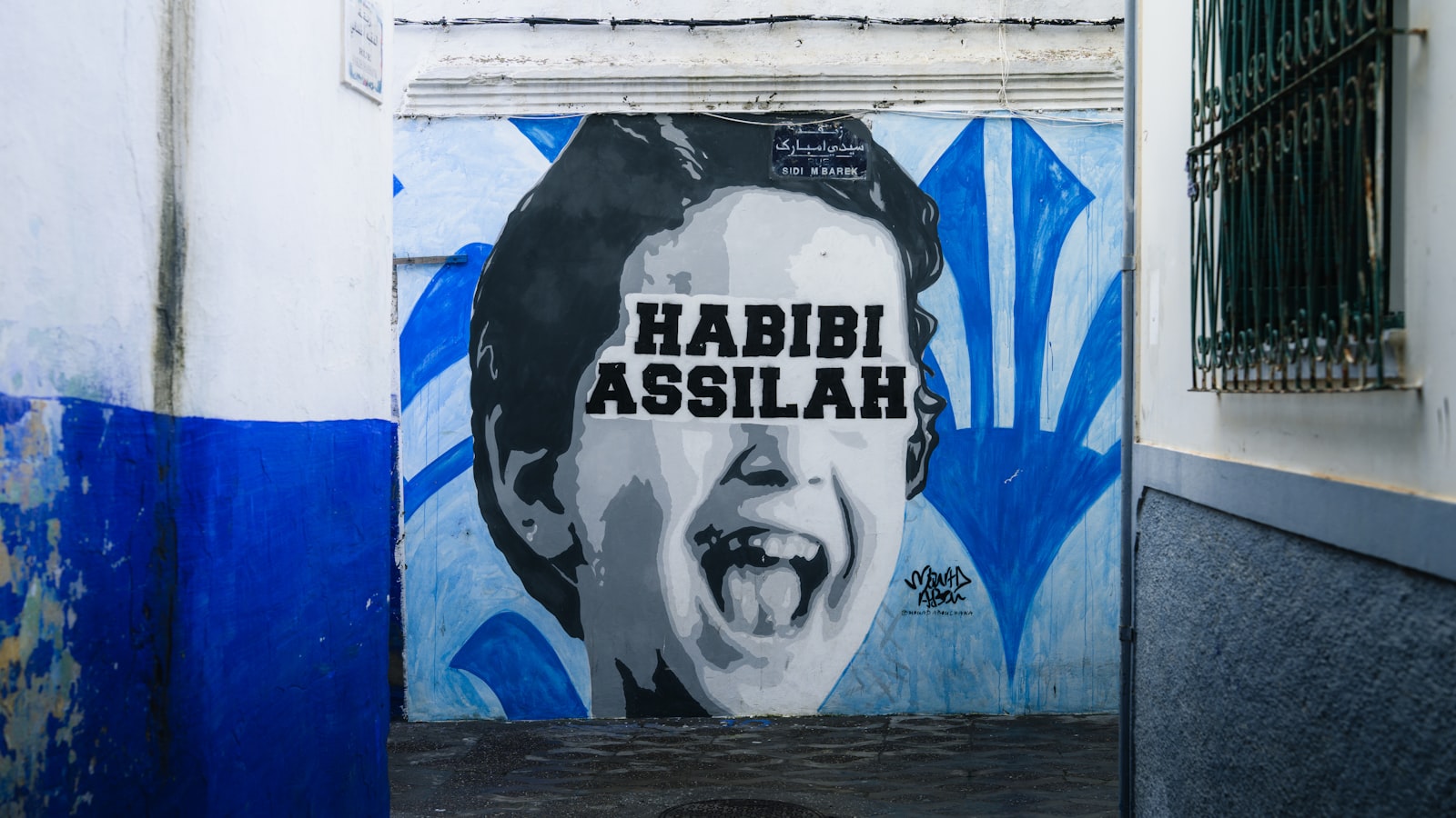 A black and white mural of a laughing child's face with the text "HABIBI ASSILAH" painted on a white wall next to a blue-painted section and a barred window.