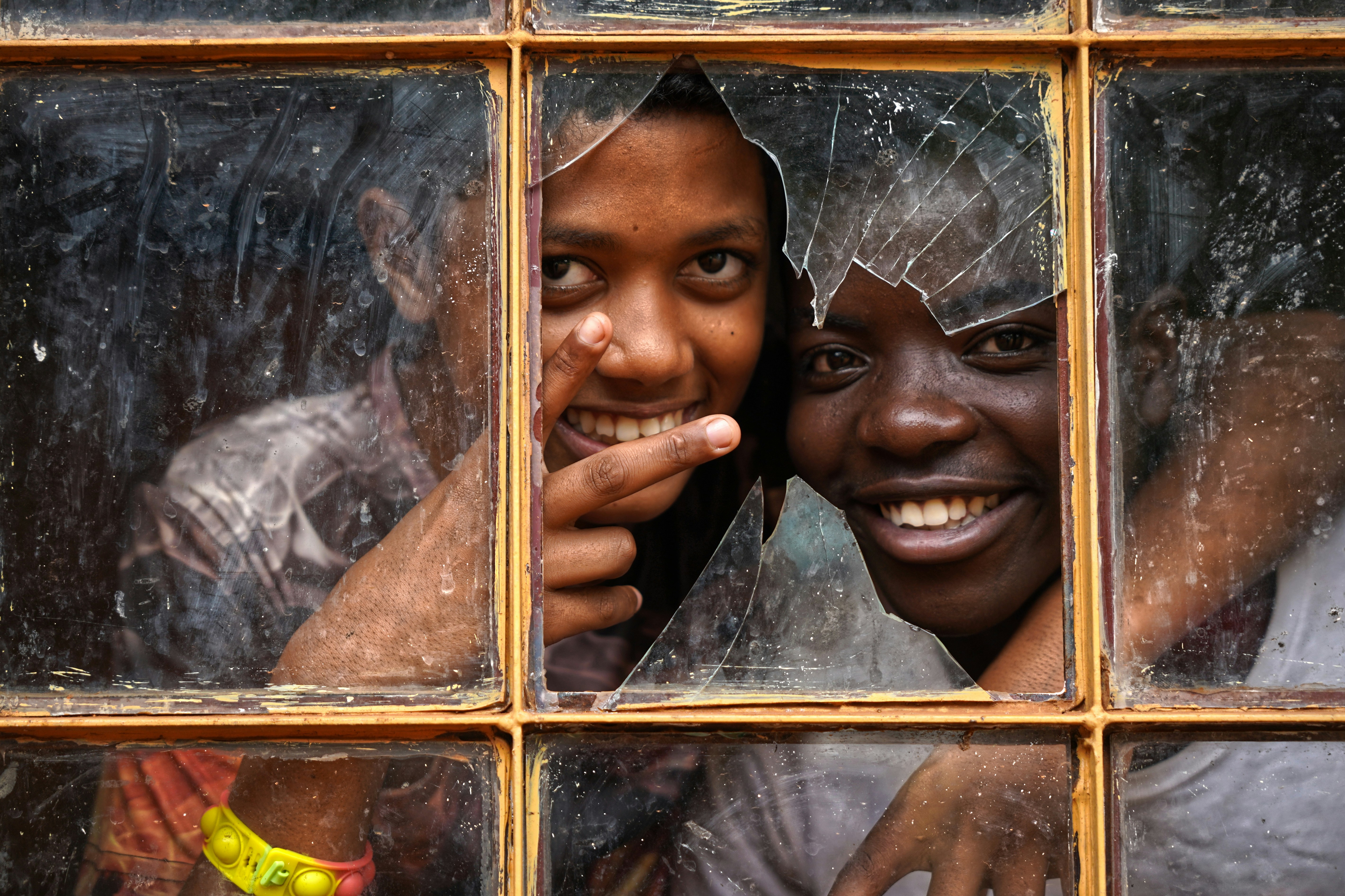 Dos niños sonrientes miran por una ventana rota.