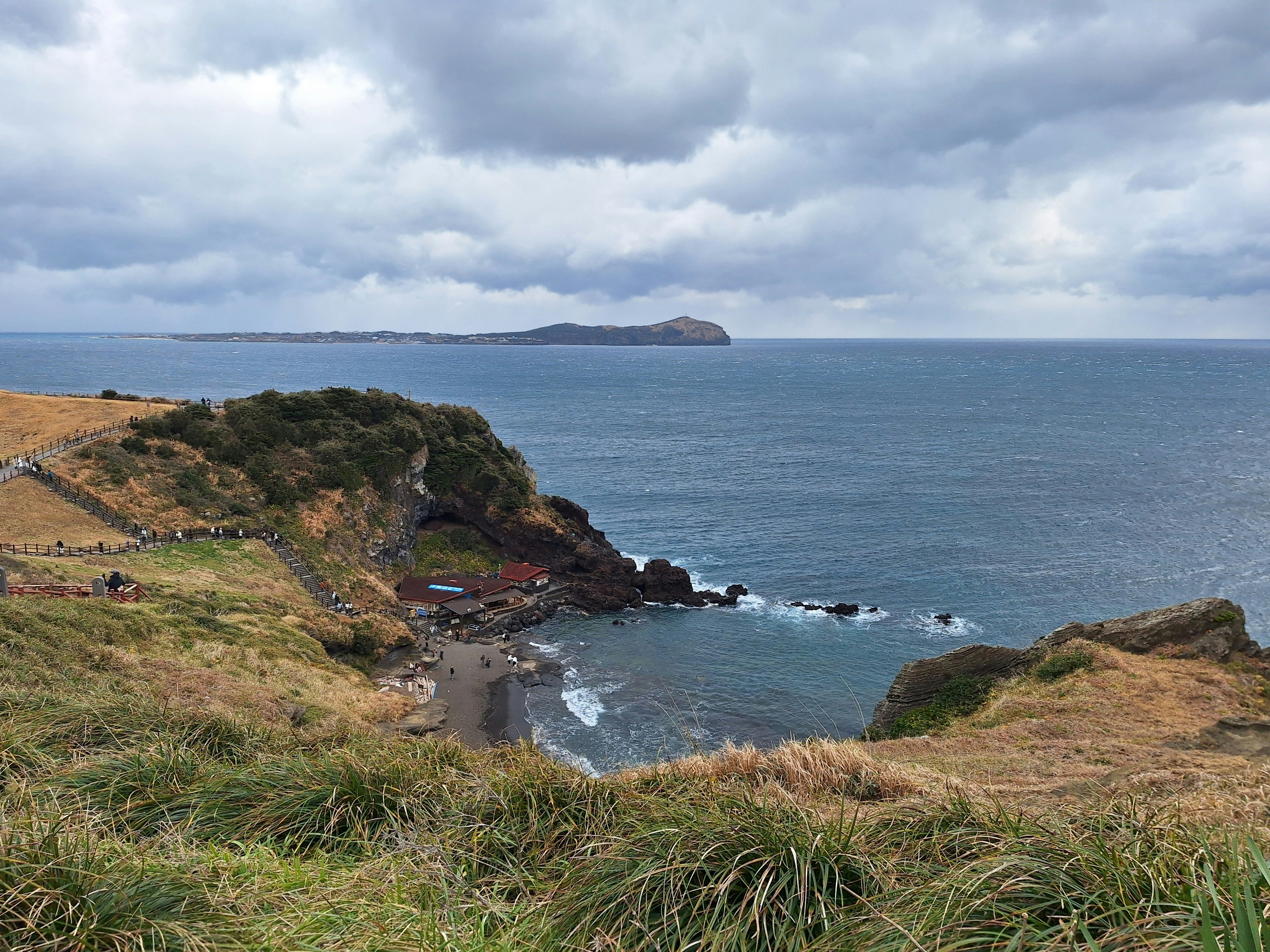 Coastal cliff overlooking a calm blue ocean under cloudy skies.