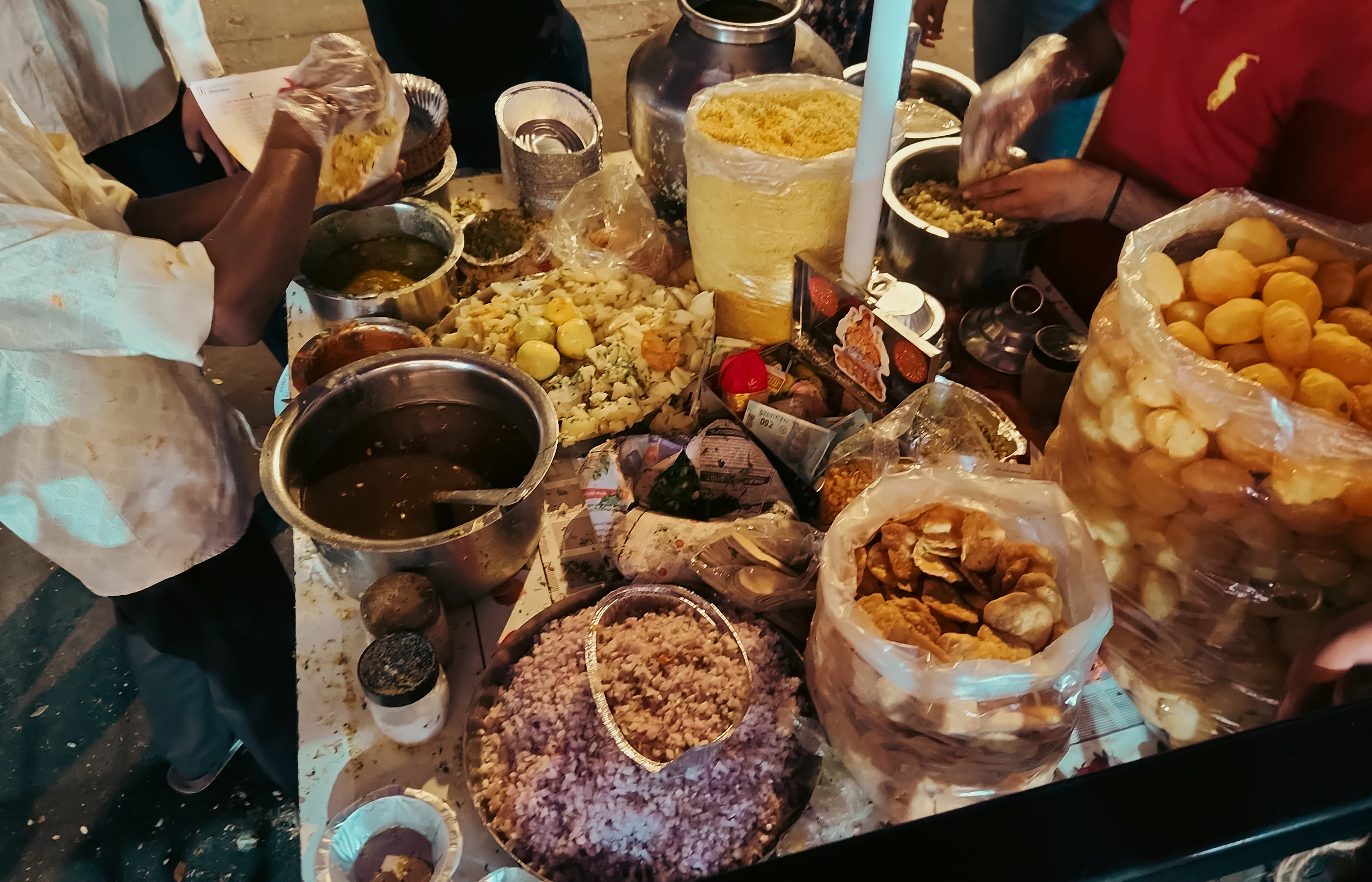 A Mumbai chaat counter in full swing: trays of crisp puris, sev, chopped onion, boiled potato and dark chutneys crowd the stall table while the vendor's hands assemble a plate.