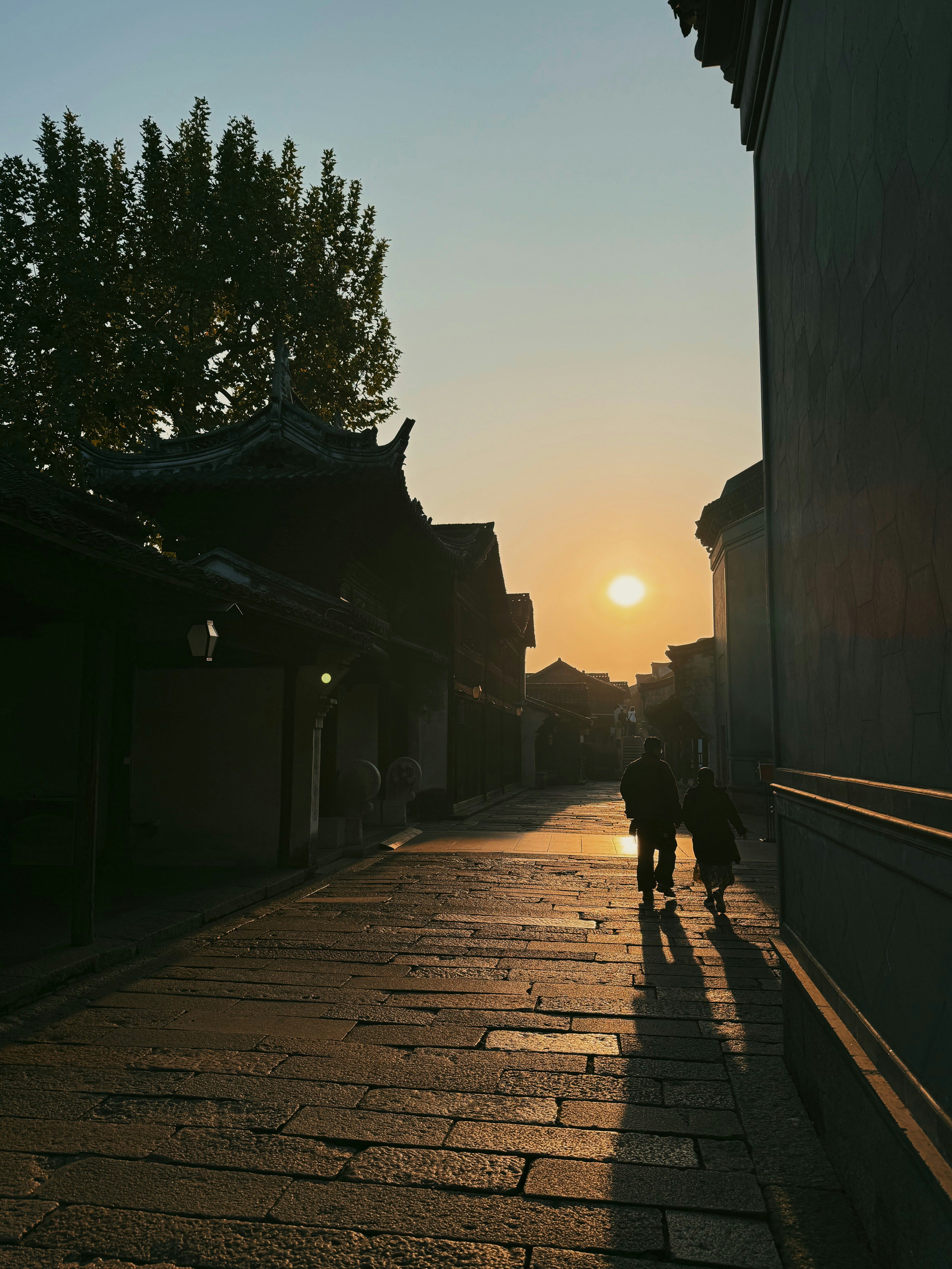 Two people walk down a cobblestone street at sunset.