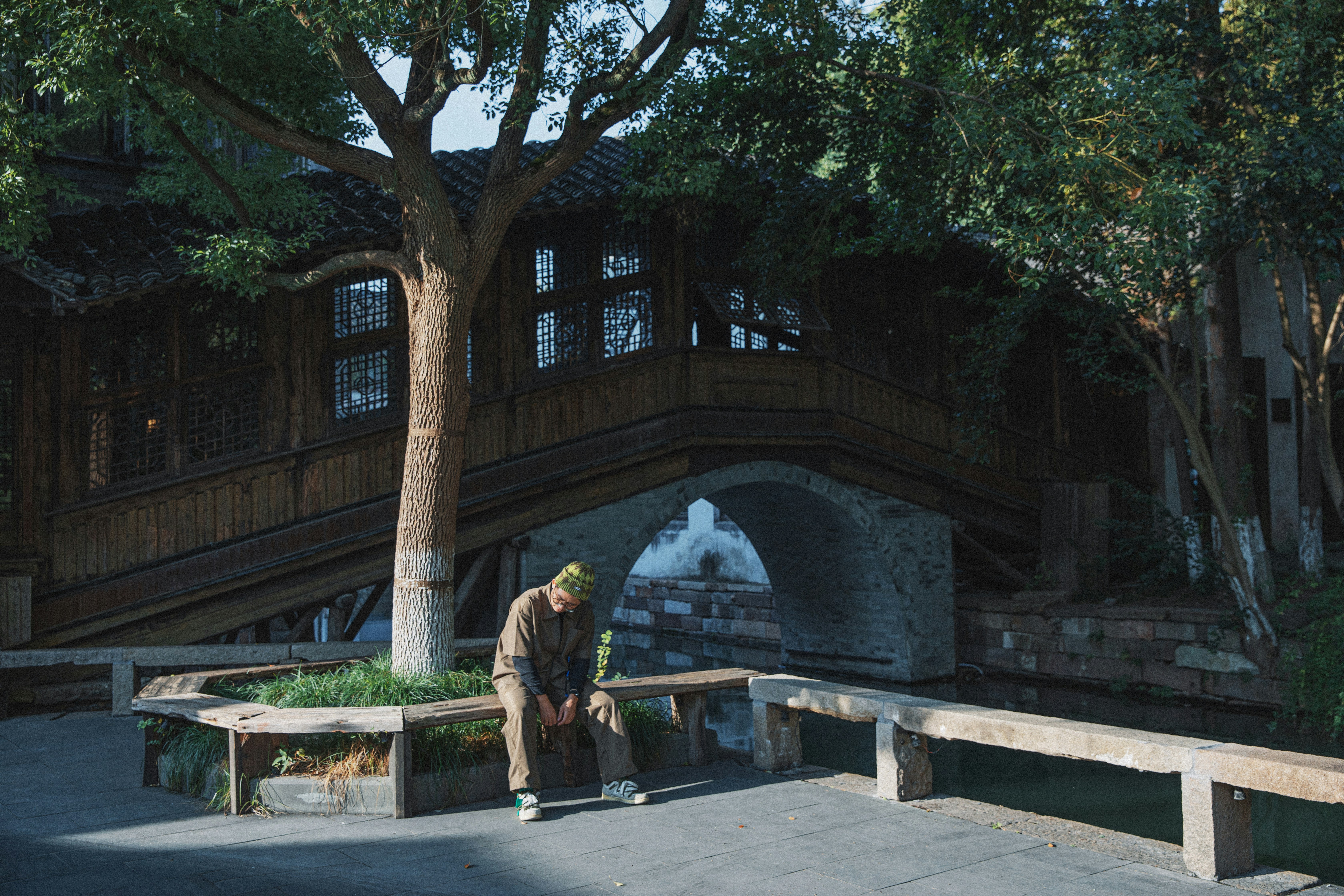 Man sits on bench near old wooden bridge and trees.