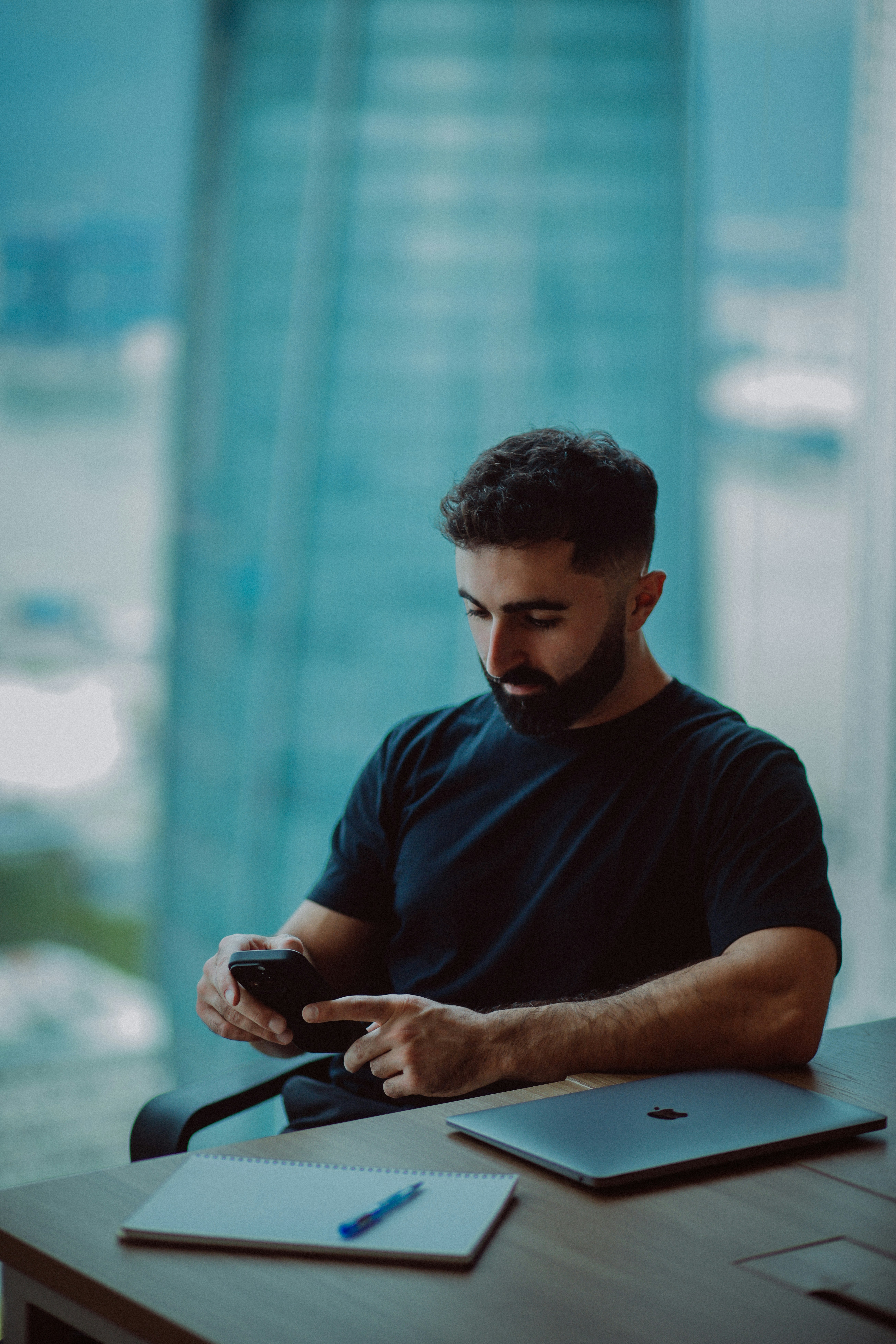 Man in black shirt using smartphone at desk with laptop.