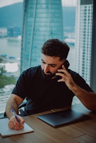 Man on phone writing in notebook at desk.