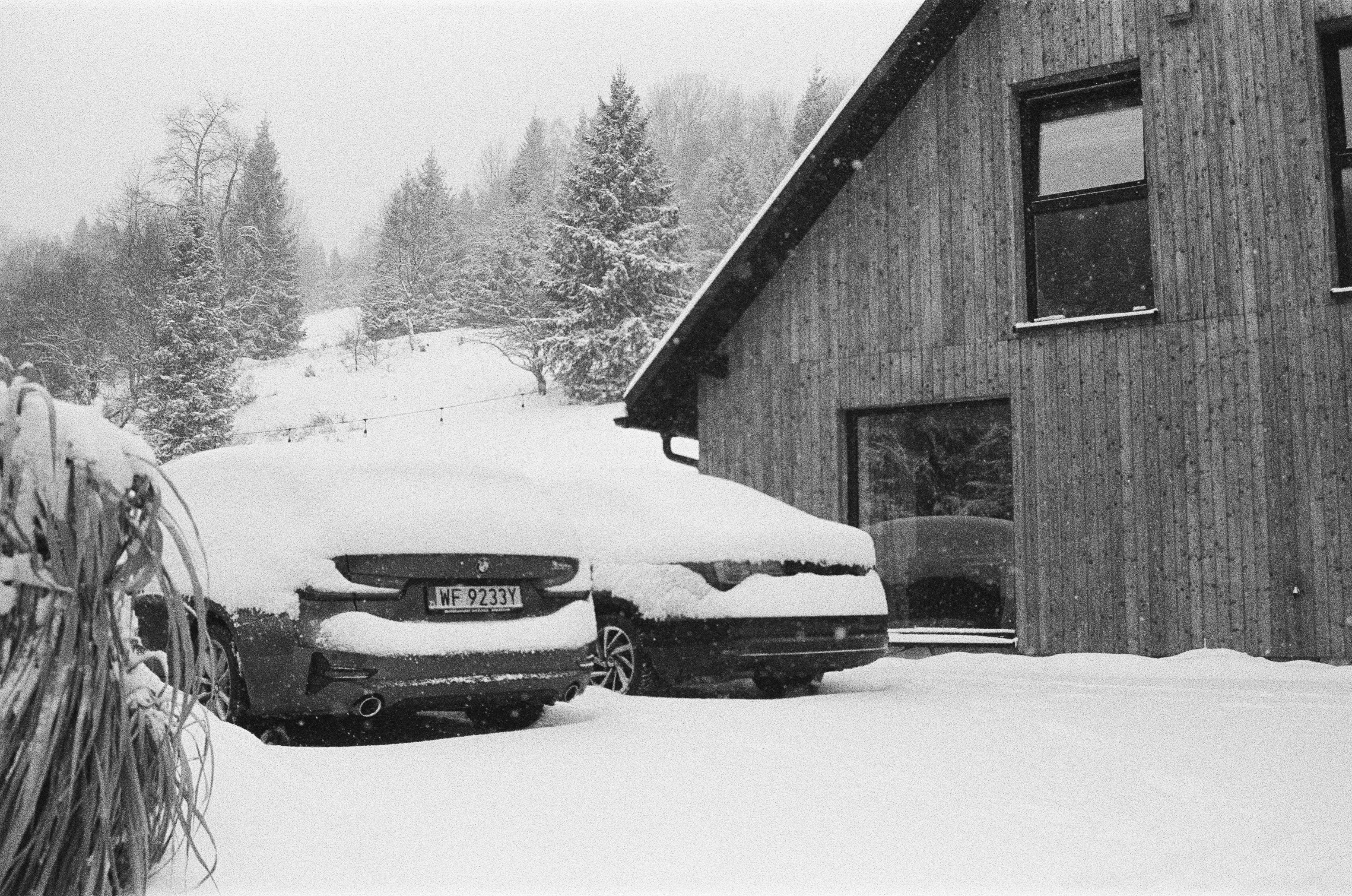 Two cars covered in snow outside a wooden house.