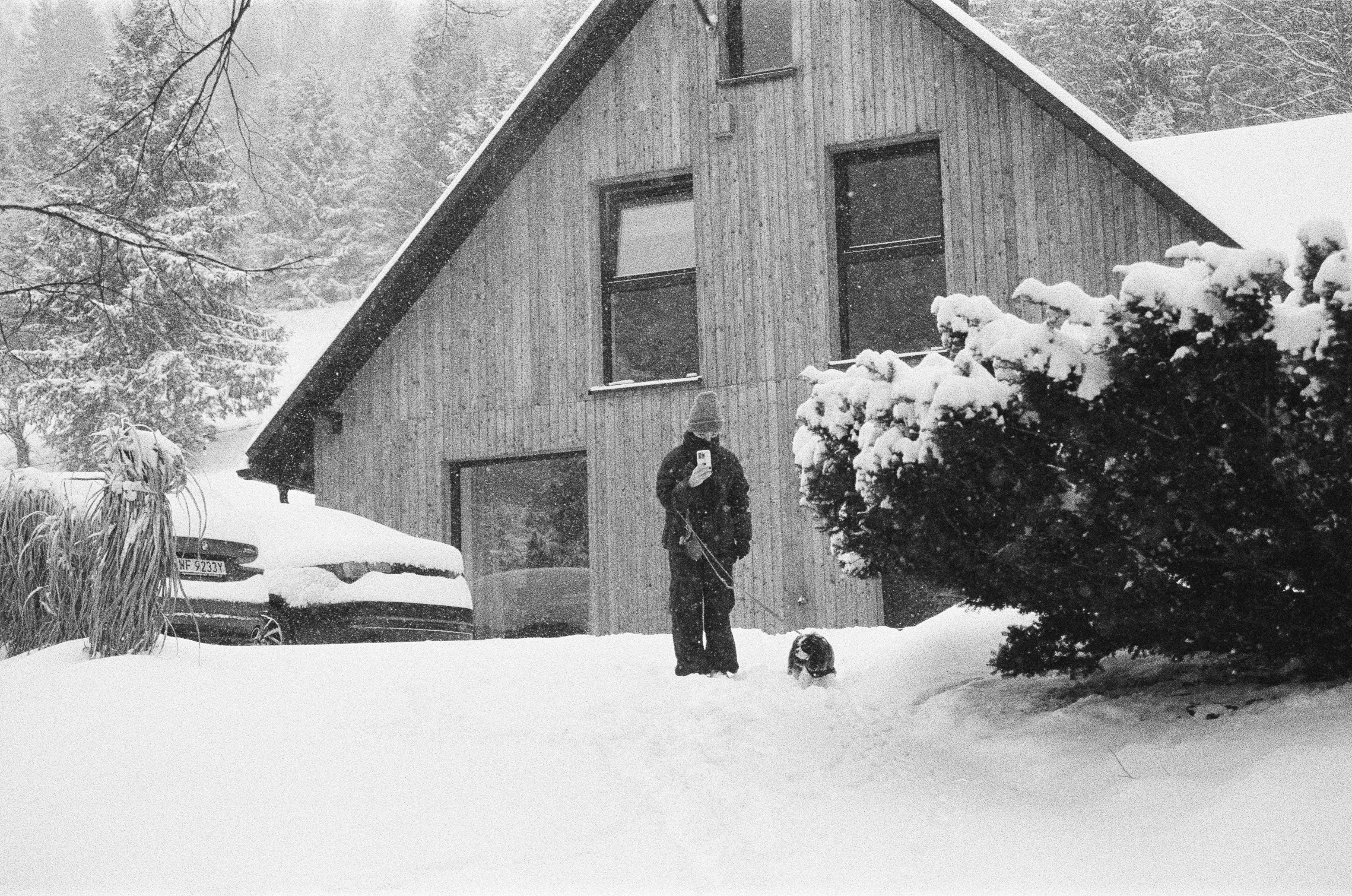 A person stands outside a wooden house in the snow.