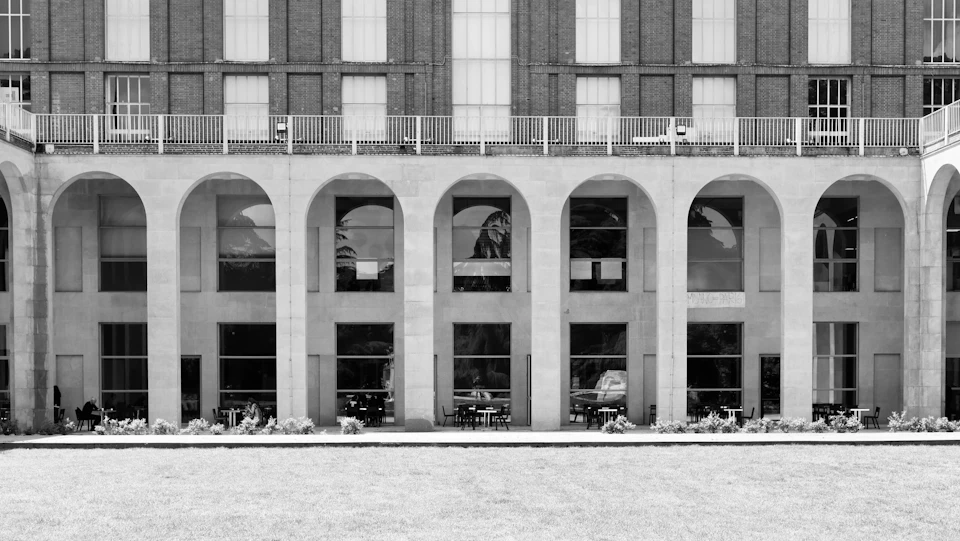 The image depicts a large, ornate building with multiple arched windows and balconies on all sides. The building is constructed from stone or concrete and features a series of arches that create an impressive architectural design. The perspective of the photo is taken from below, looking up at the building's facade, emphasizing its grandeur and imposing presence. In front of the building, there are several potted plants scattered around, adding a touch of greenery to the scene.