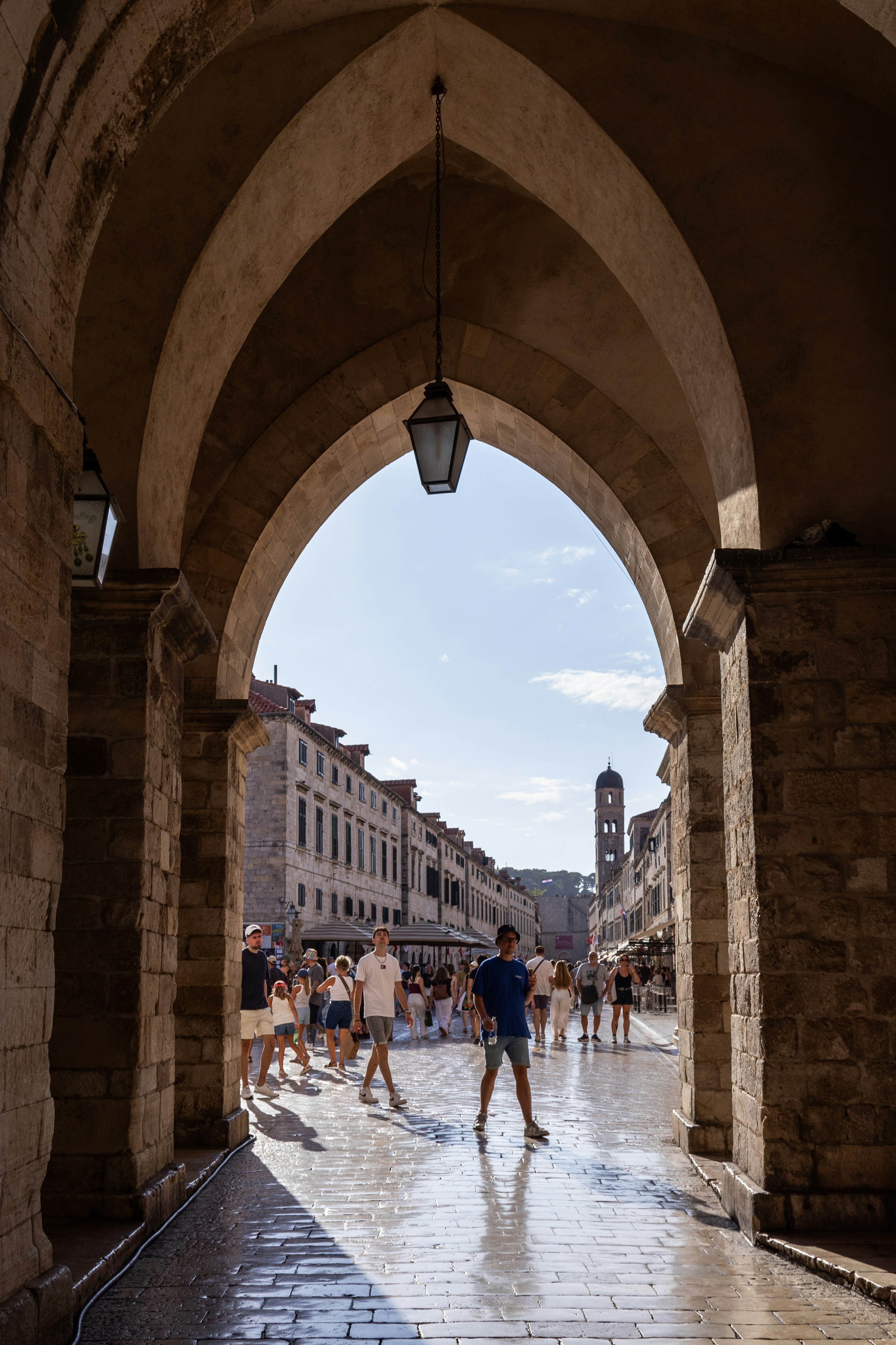 People walk through stone archway onto sunny cobblestone street.