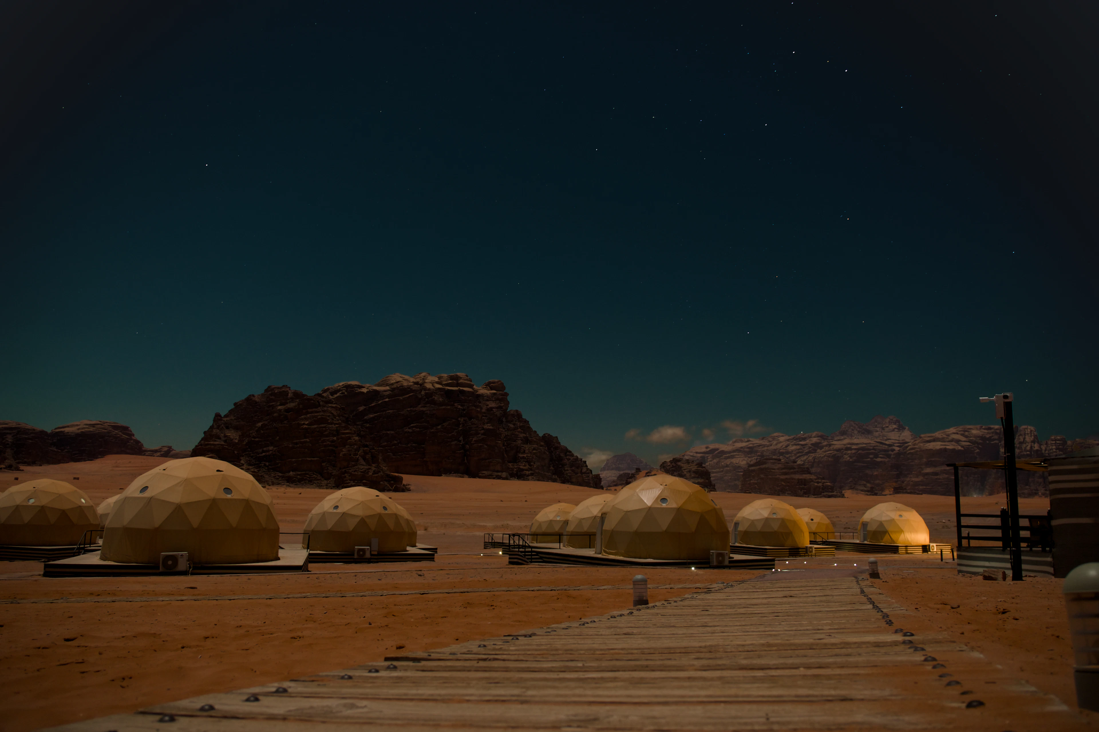 A row of geodesic dome tents glowing amber on the red sand of Wadi Rum at night, a lit boardwalk running between them and stars above the dark sandstone mesas behind.