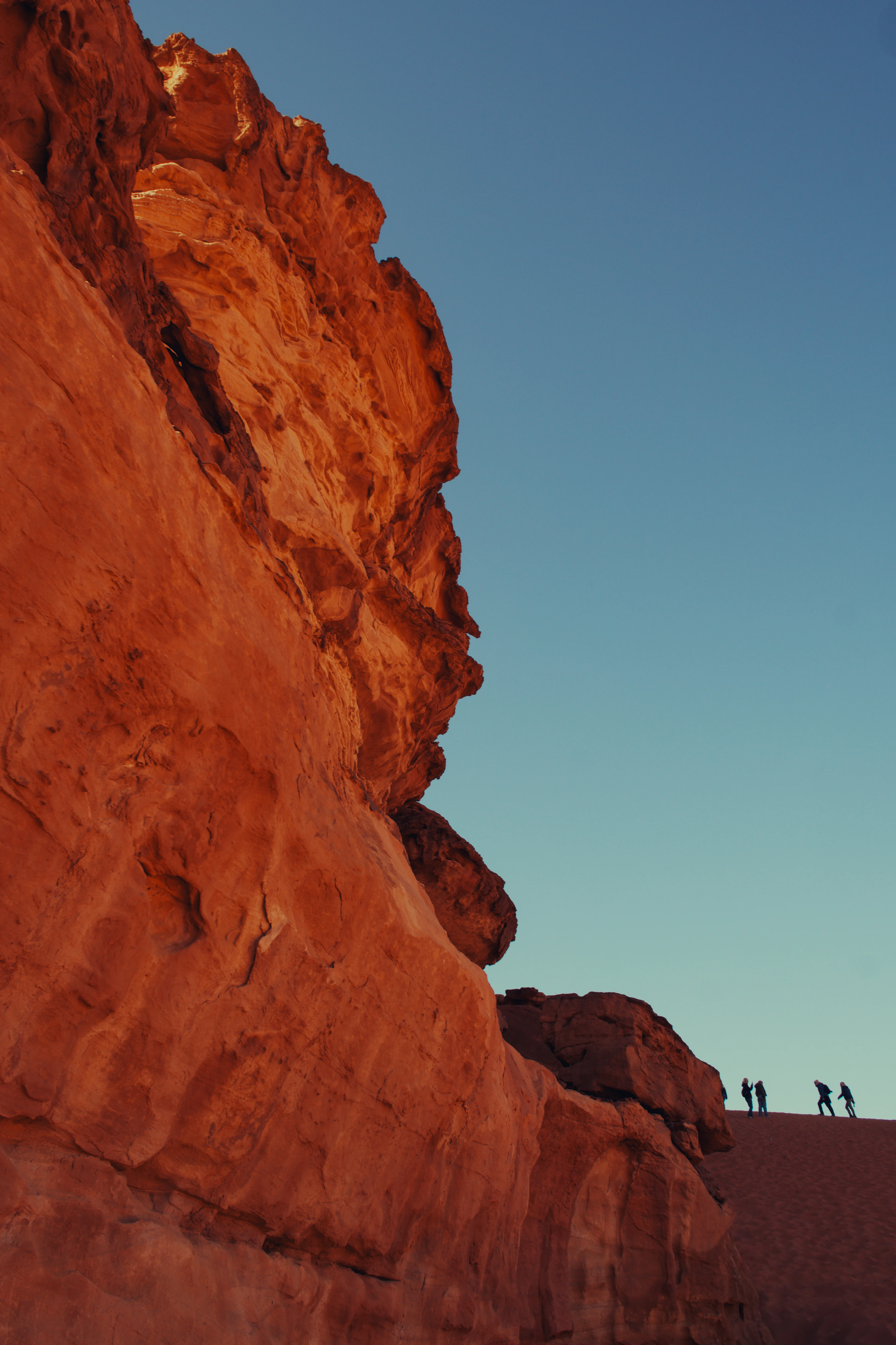 A sheer red sandstone buttress in Wadi Rum glowing in late light, with four tiny walkers silhouetted on the sand ridge beside it to show its scale.