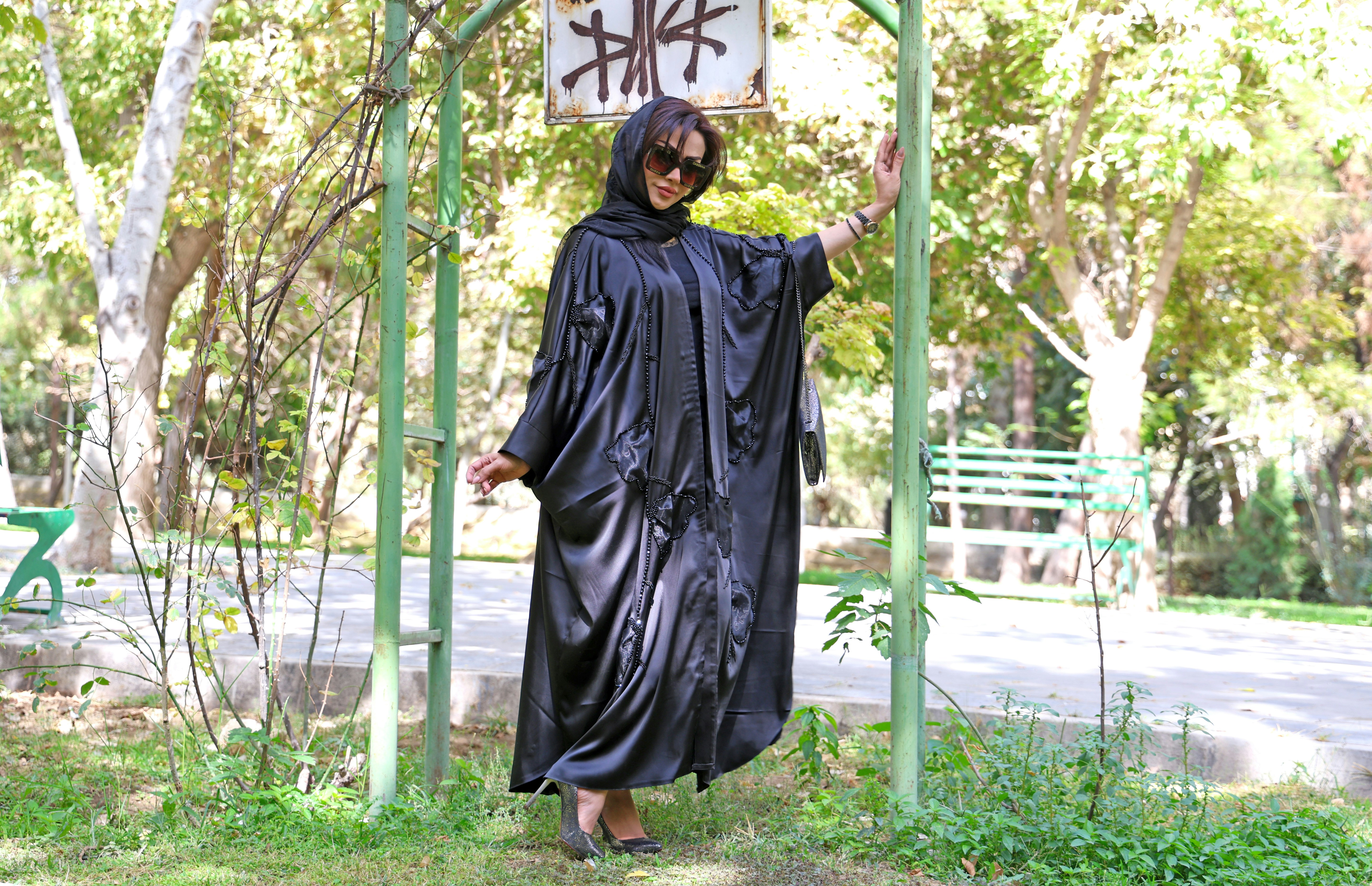 Wanita berbaju abaya hitam berdiri di luar ruangan dekat pepohonan.