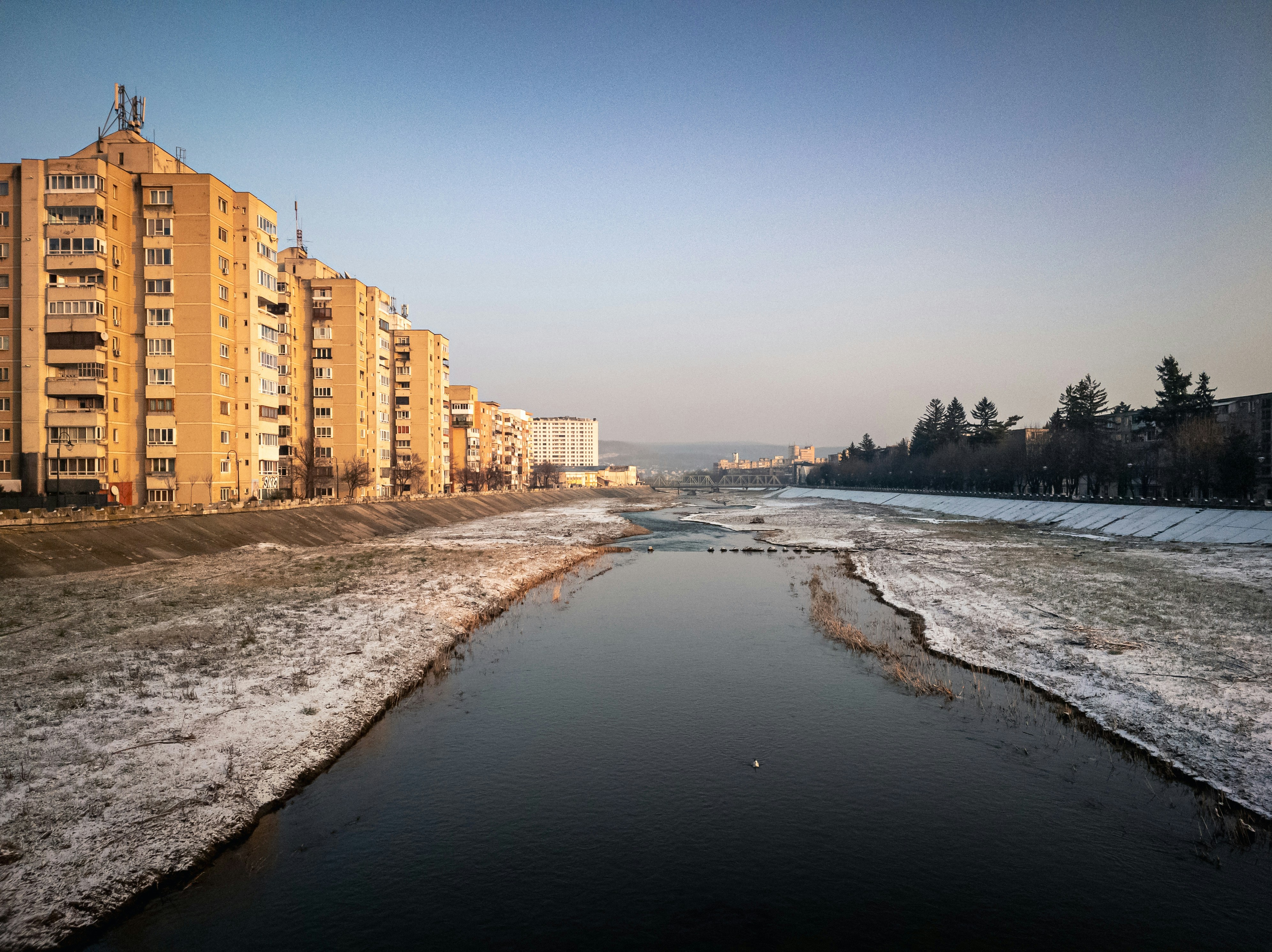 Apartment buildings line a river with snow on the banks.