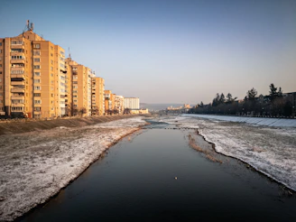 Apartment buildings line a river with snow on the banks