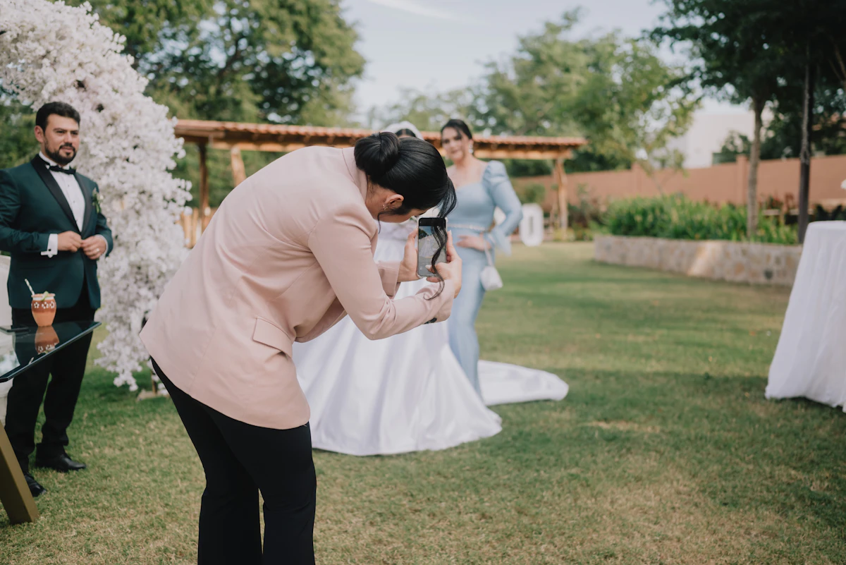 A photographer in a blush blazer leans in to capture a bride on her phone at a garden wedding as the groom looks on