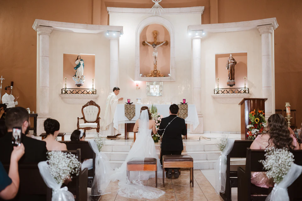 A bride and groom at the altar of a church during a Catholic wedding ceremony, with white flowers tied to the pew ends